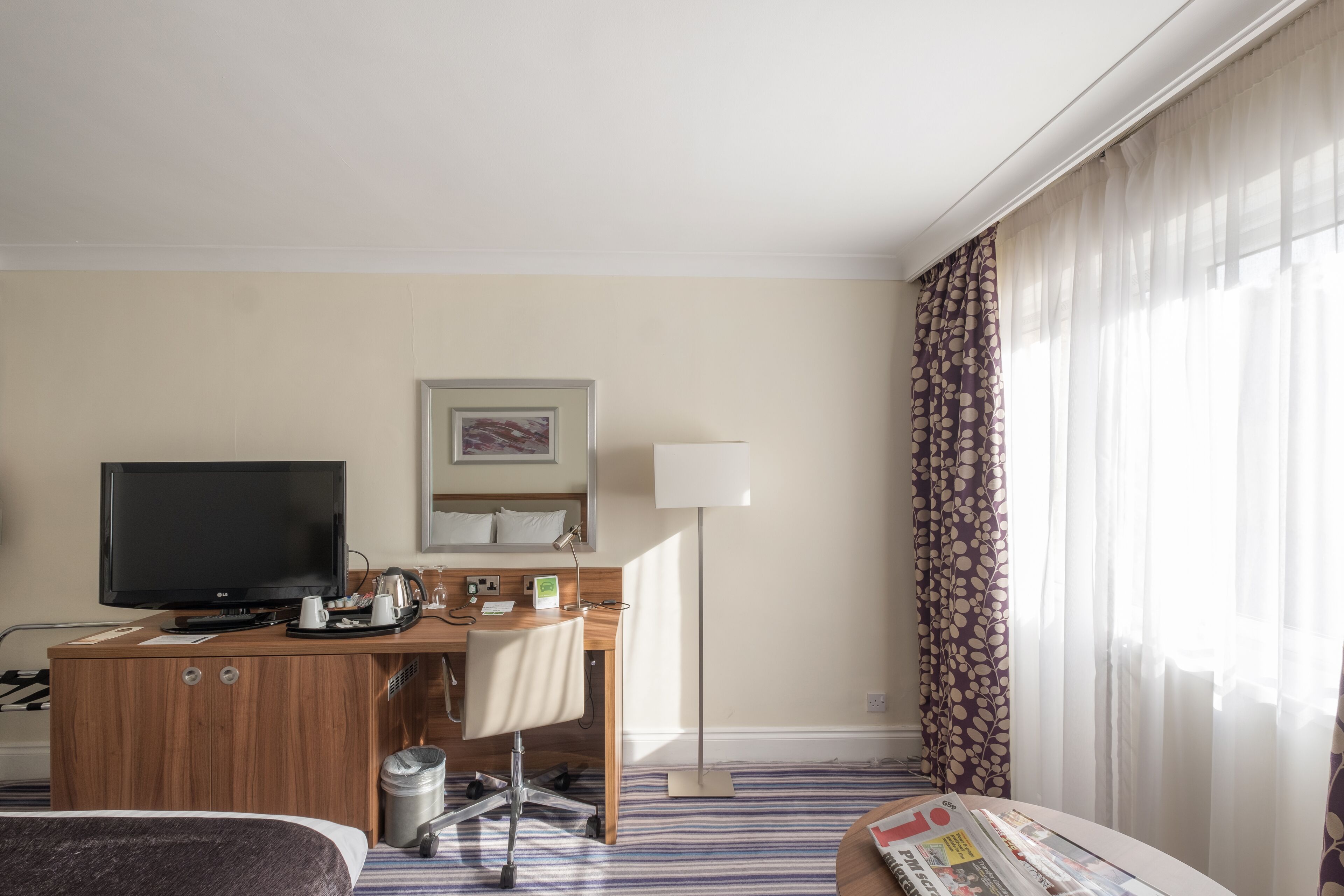 A hotel room with a bed, a desk with a computer, a television, and a window with curtains.