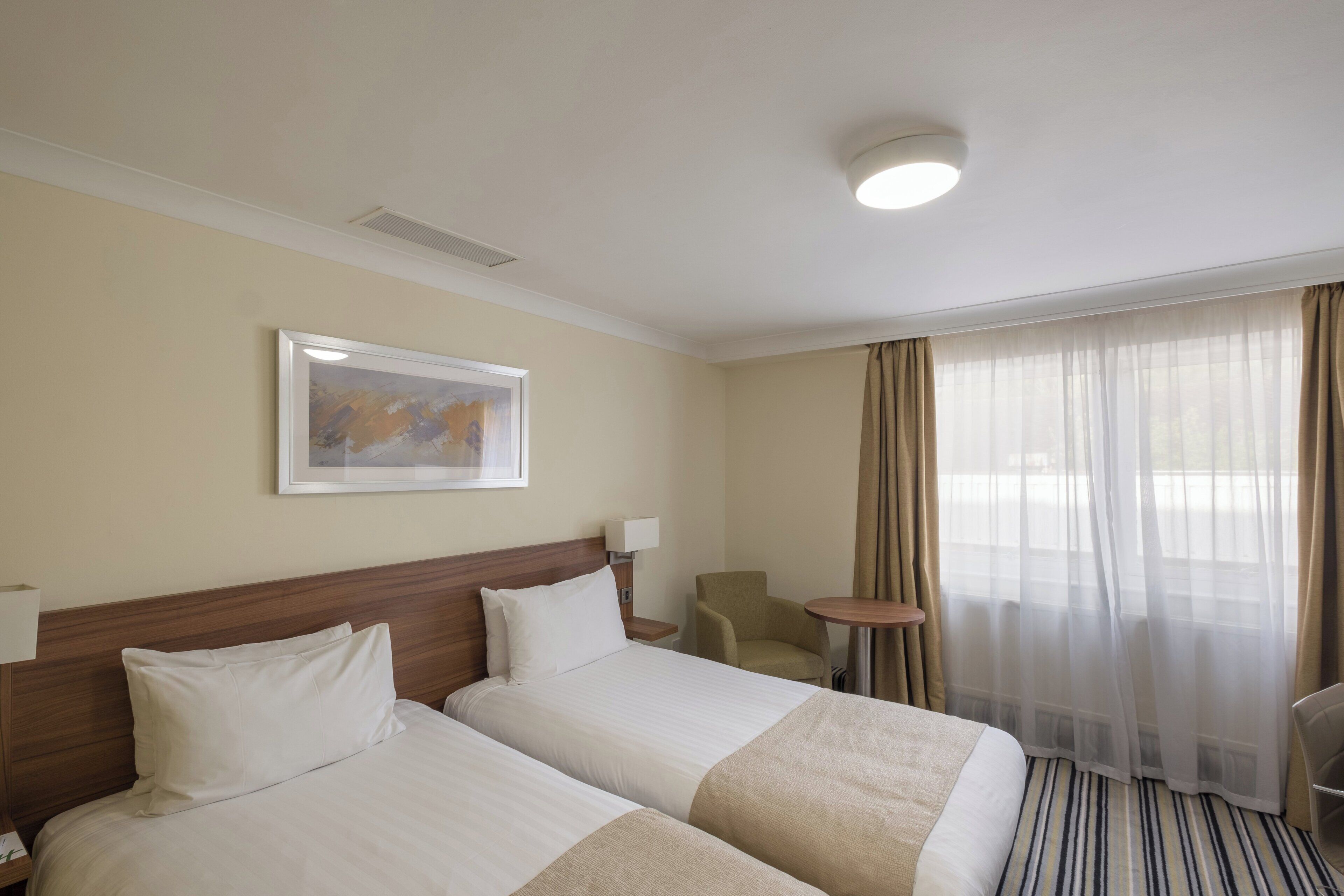 A hotel room with two beds, a headboard, a framed picture, a lamp, a chair, a small table, and a window with curtains.