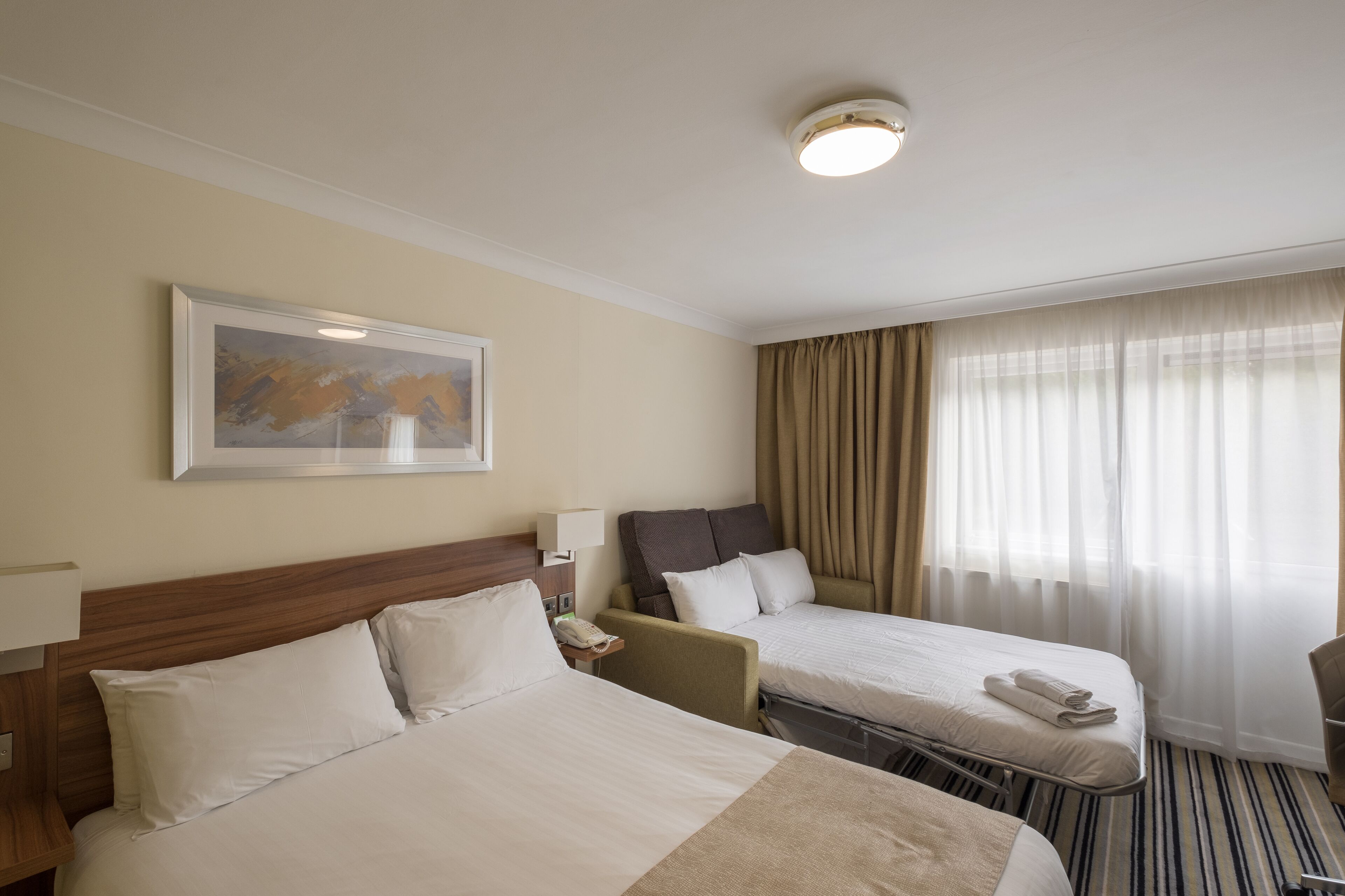 A hotel room with two beds, a painting on the wall, a lamp, and a window with curtains.