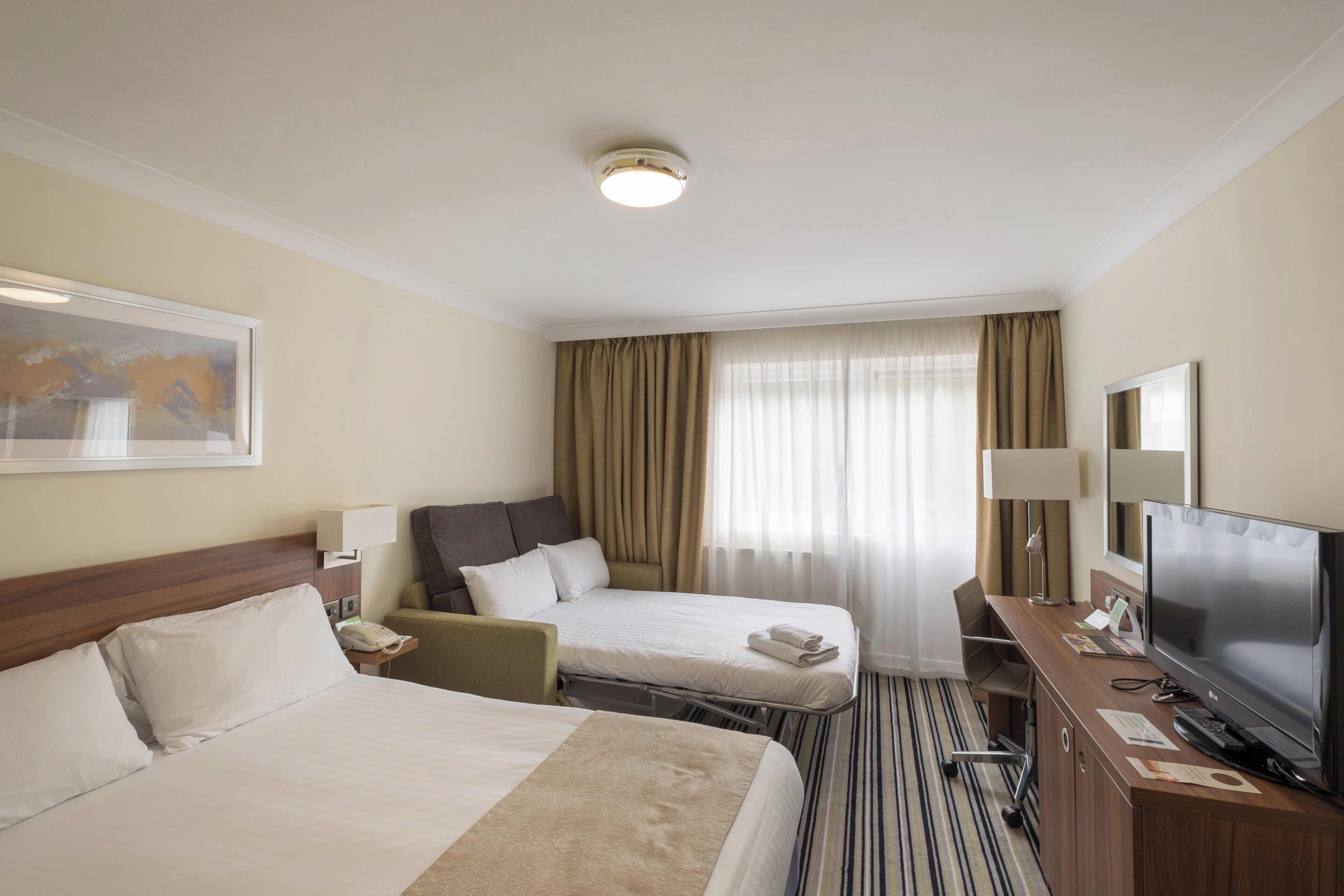 A hotel room with two beds, a desk with a television, a chair, a lamp, and a window with curtains.