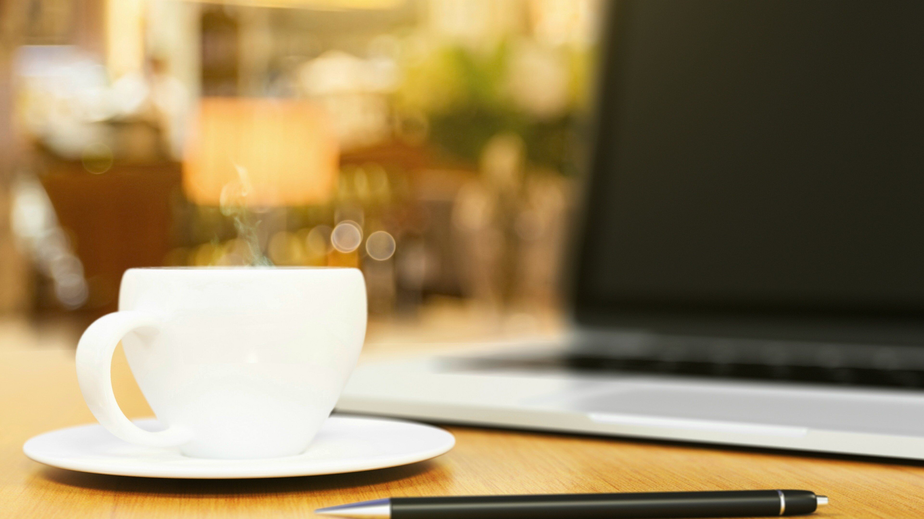 A white cup on a saucer with steam rising, a black pen, and a laptop on a wooden surface.