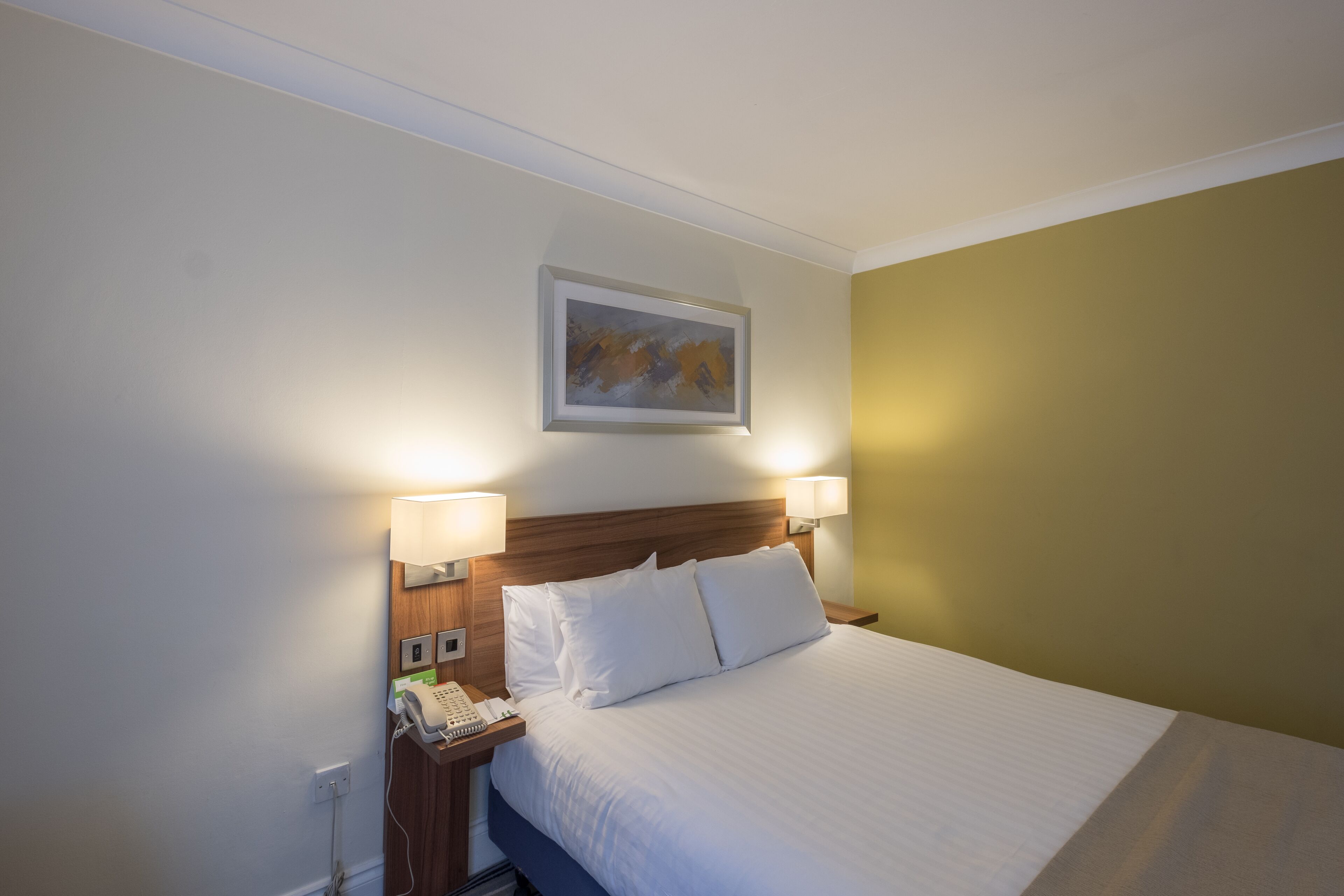 A hotel room with a neatly made bed, two bedside lamps, a telephone, and a framed artwork on the wall.