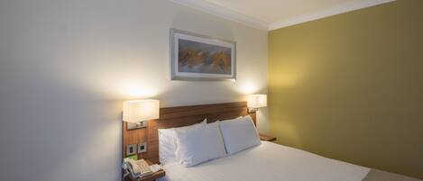 A hotel room with a neatly made bed, two bedside lamps, a telephone, and a framed artwork on the wall.