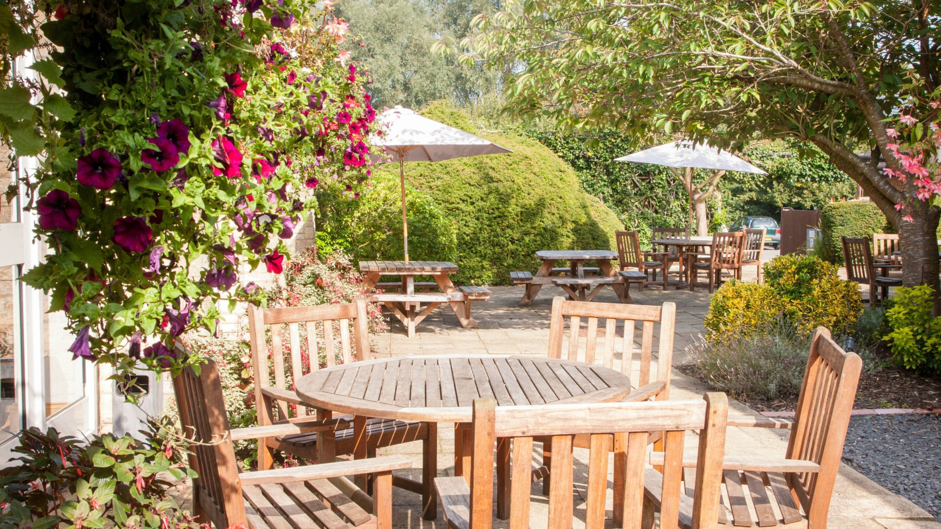 An outdoor patio with wooden tables and chairs, surrounded by lush greenery and flowering plants.