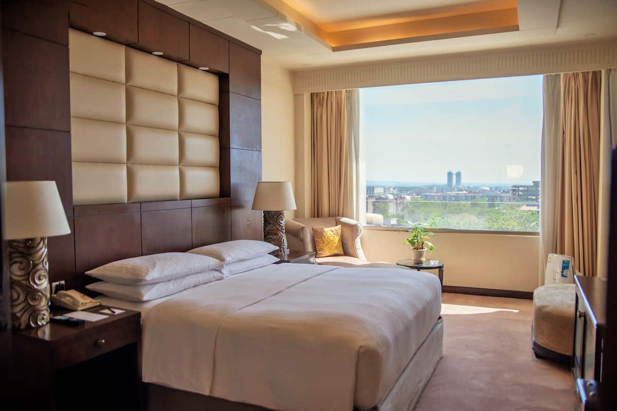 Habitación de hotel con una cama grande, dos mesitas de noche con lámparas, una mesita auxiliar con una planta y vistas de la ciudad a través de un ventanal amplio.