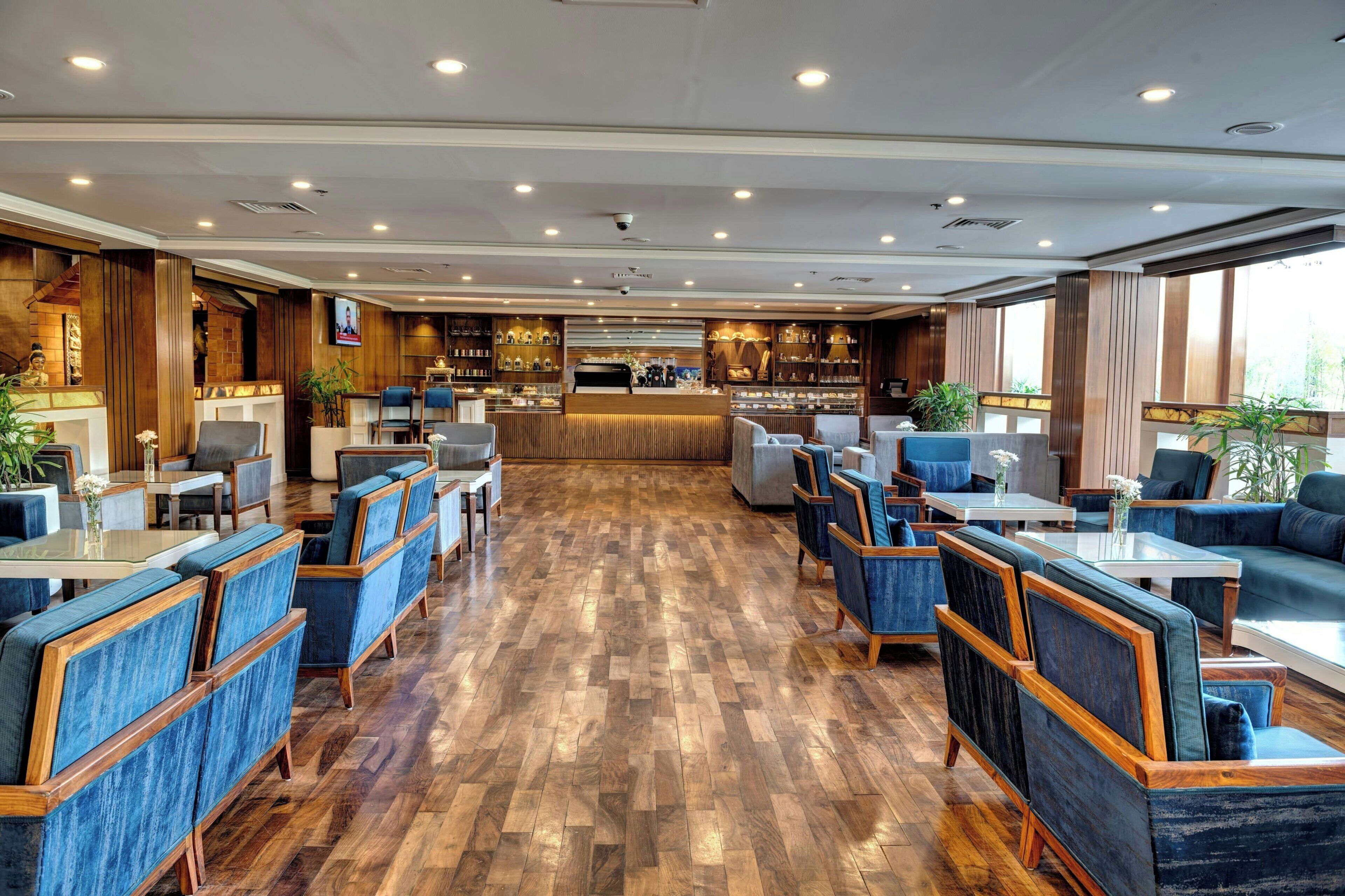 Un hall d’hôtel spacieux, doté d’un plancher en bois, de sièges rembourrés en tissu bleu, d’un bar et de grandes fenêtres.