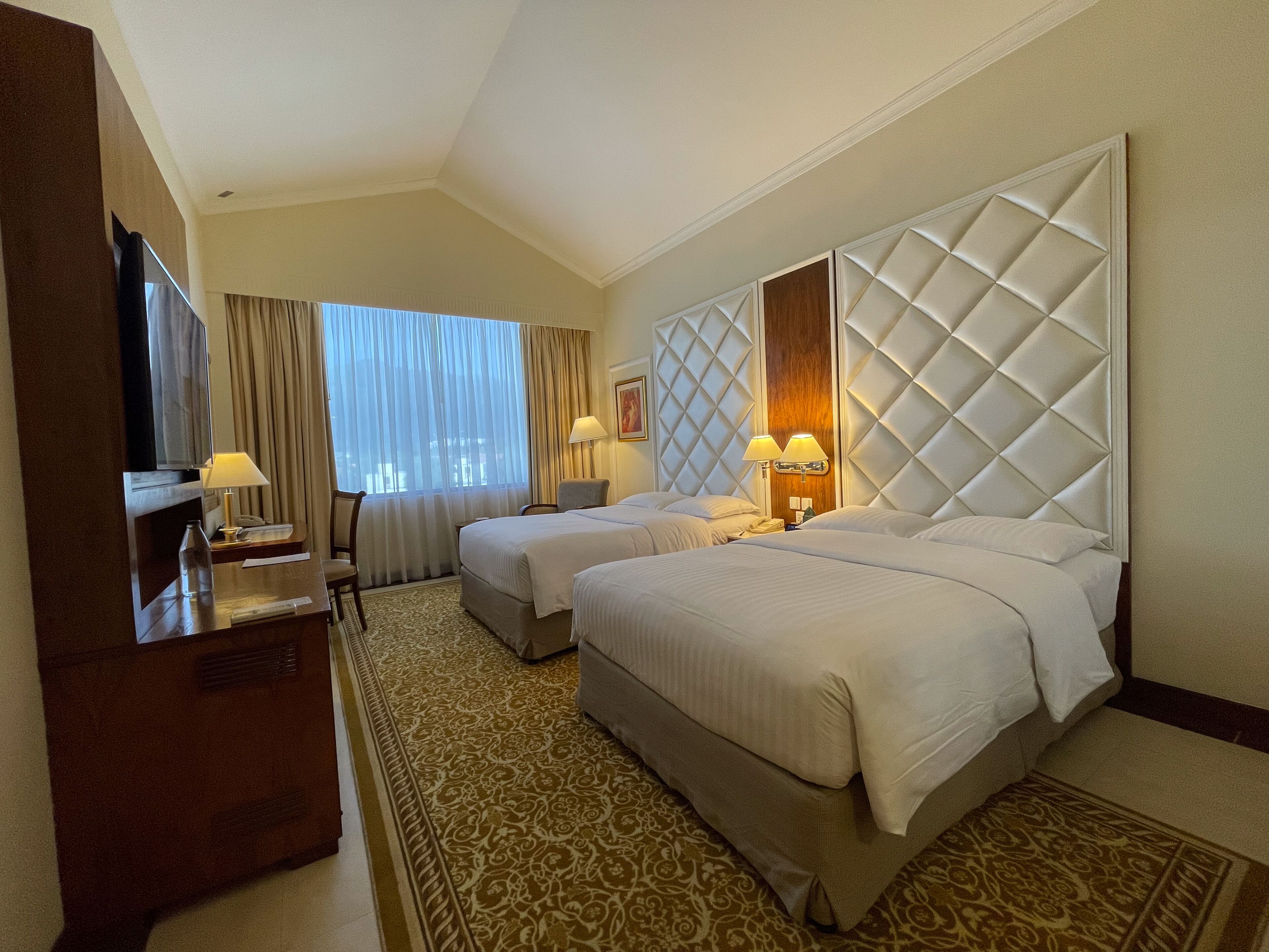 A hotel room with two beds, a desk, a TV, and a large window with curtains.