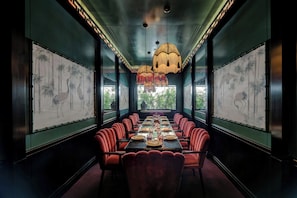 A long dining table with red chairs, flanked by large windows with bamboo artwork.