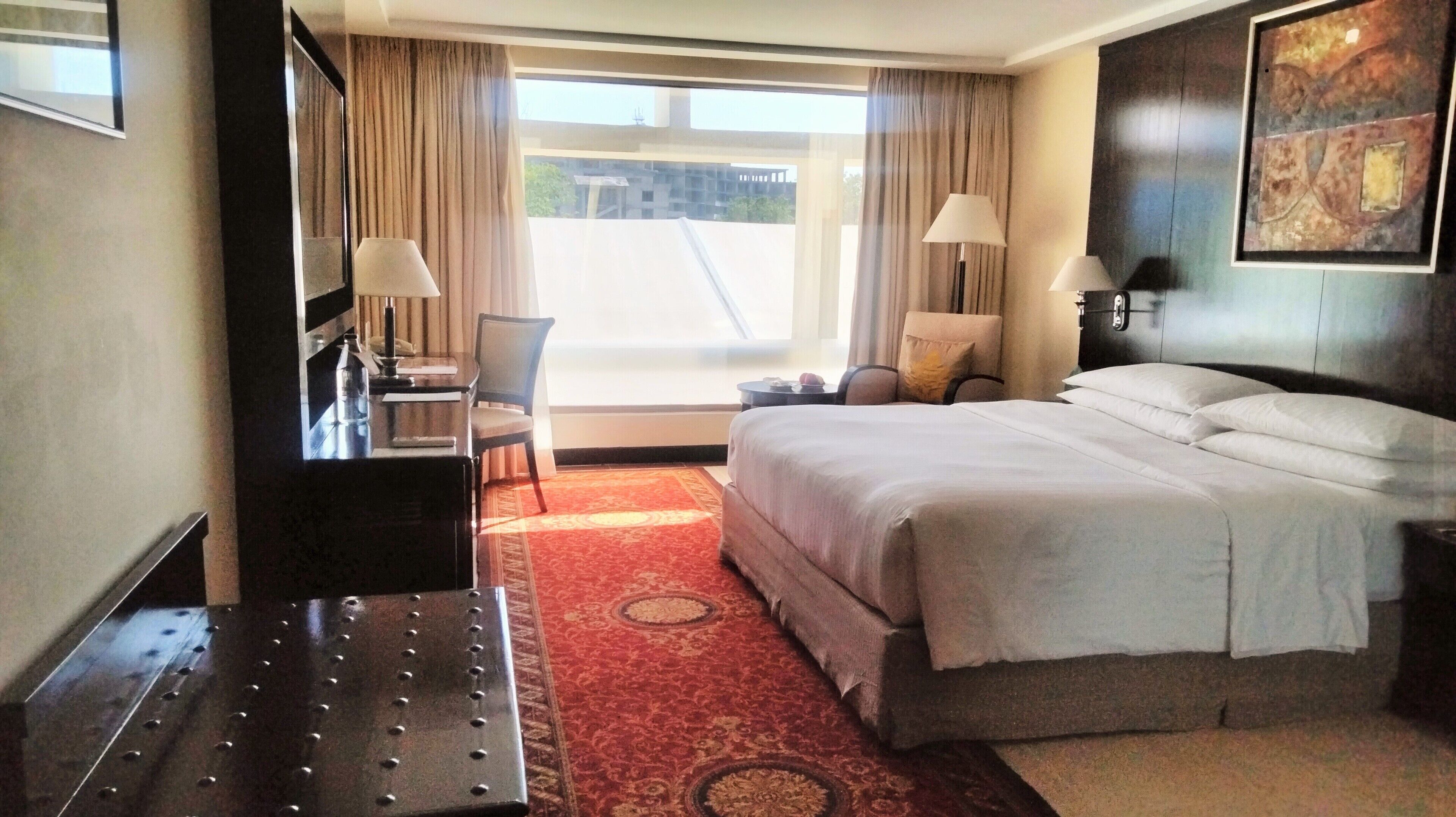 Een hotelkamer met een bed, twee stoelen, een bureau, een lamp en een groot raam met gordijnen.