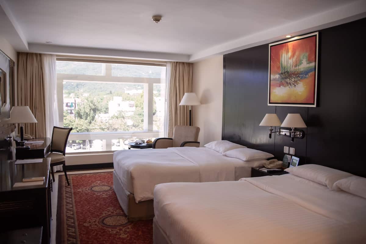 Habitación de hotel con dos camas, un escritorio, una silla, una lámpara y un cuadro en la pared.