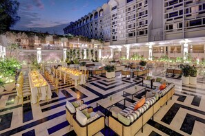 A spacious outdoor lounge area with multiple seating arrangements, including sofas and tables, surrounded by potted plants and a decorative railing.
