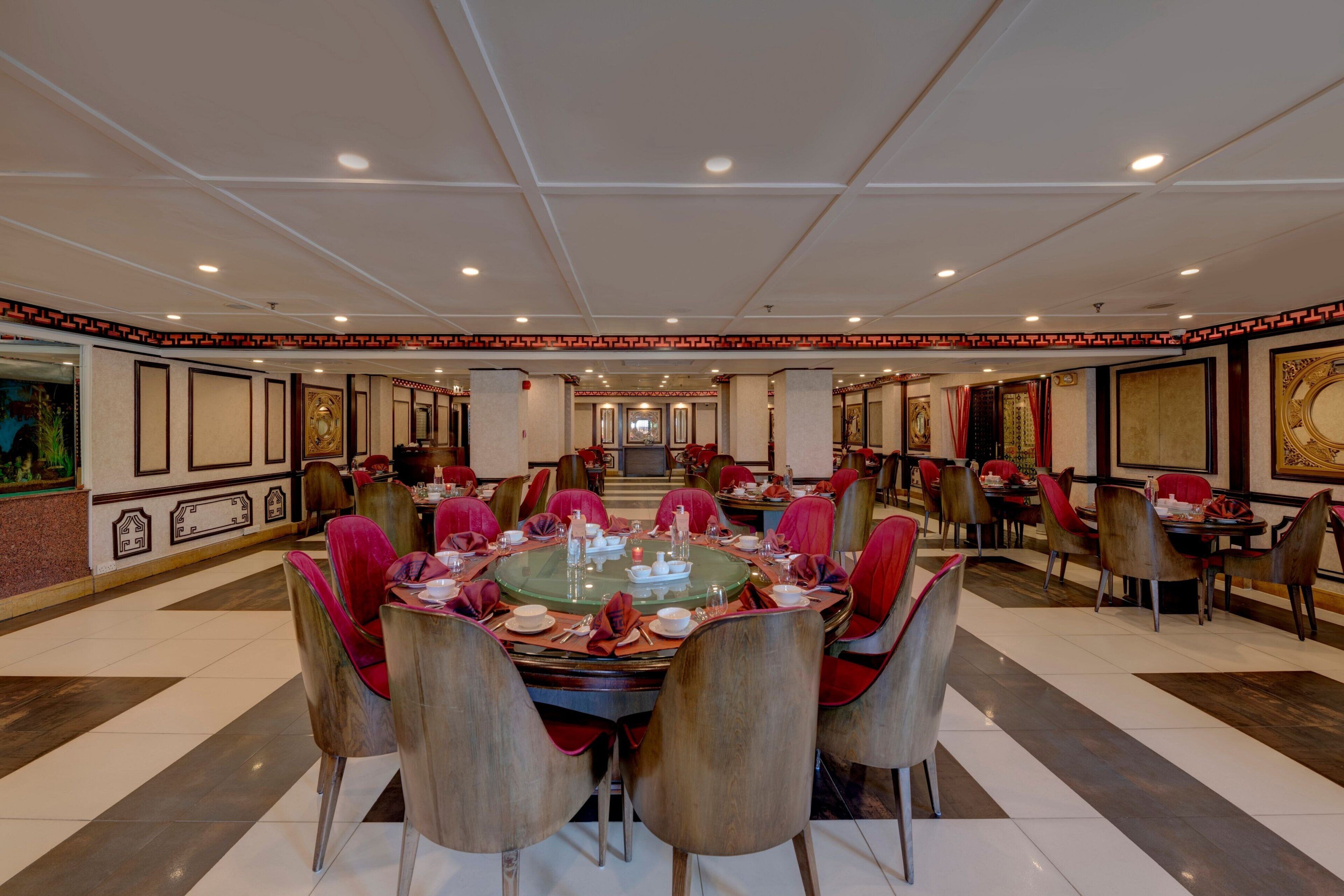 A spacious dining area with round tables and red chairs, featuring a central chandelier and framed artwork on the walls.
