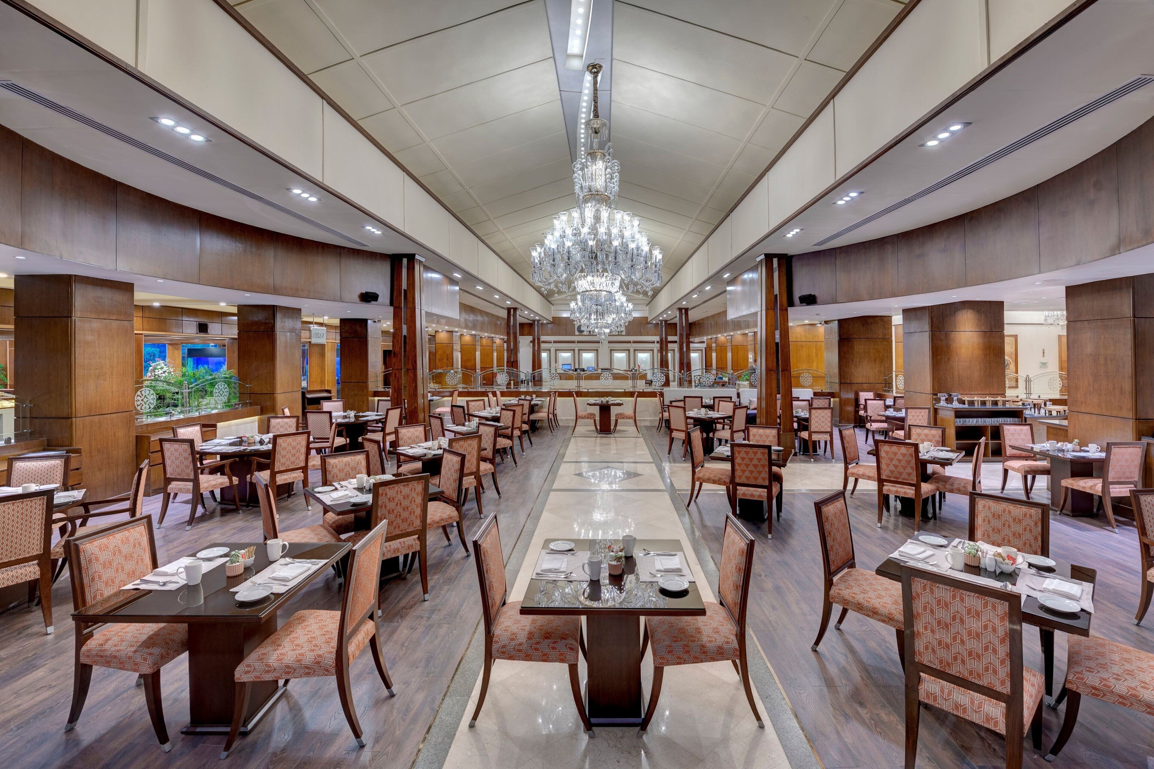 A spacious dining area with multiple tables and chairs, a large chandelier, and wooden paneling on the walls.