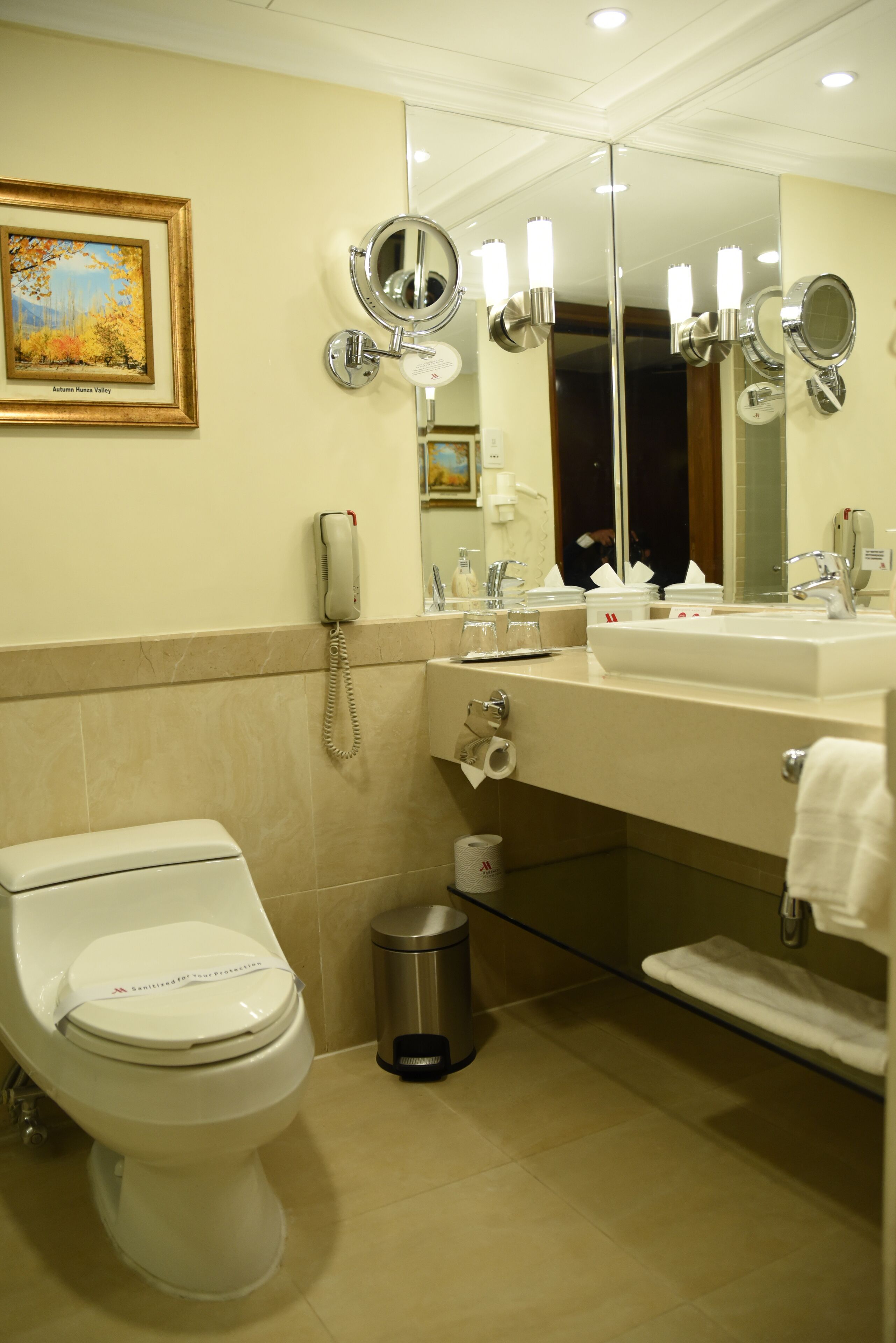 Un baño con inodoro, lavamanos y espejo. Hay un cuadro en la pared y un cubo de basura.