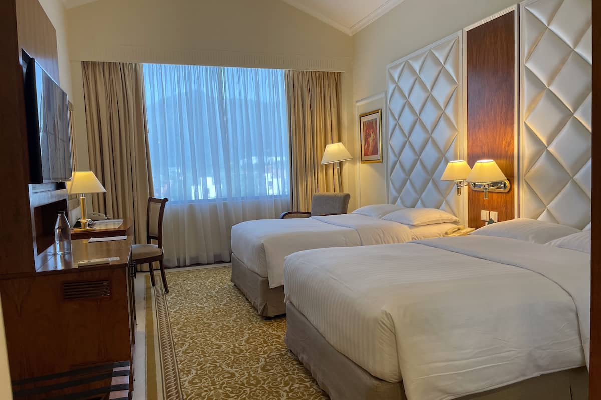 Habitación de hotel con dos camas, un escritorio con lámpara, una silla, un televisor y un ventanal con cortinas.
