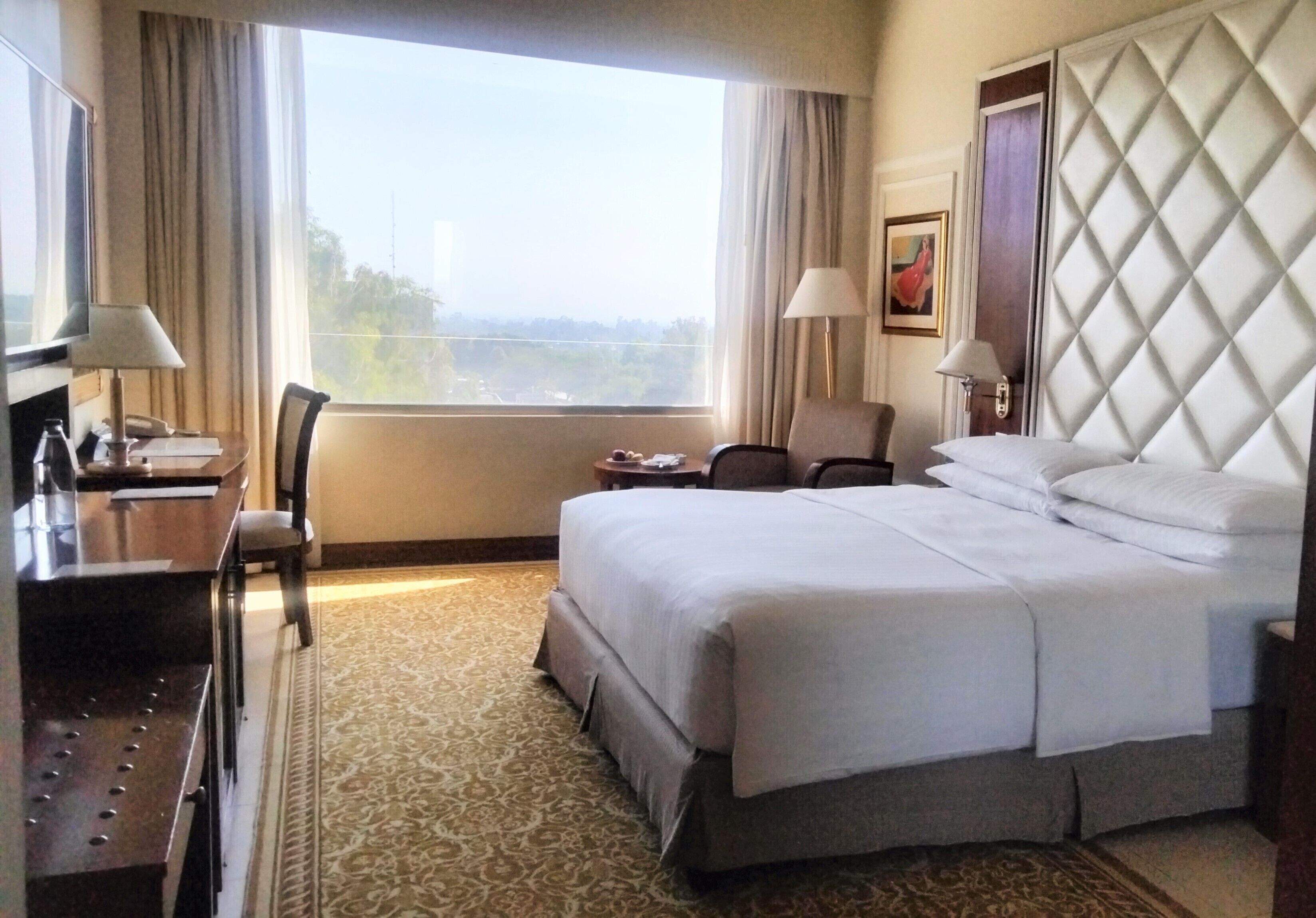 Habitación de hotel con una cama grande, un escritorio con lámpara, una silla y vista a la ciudad por la ventana.