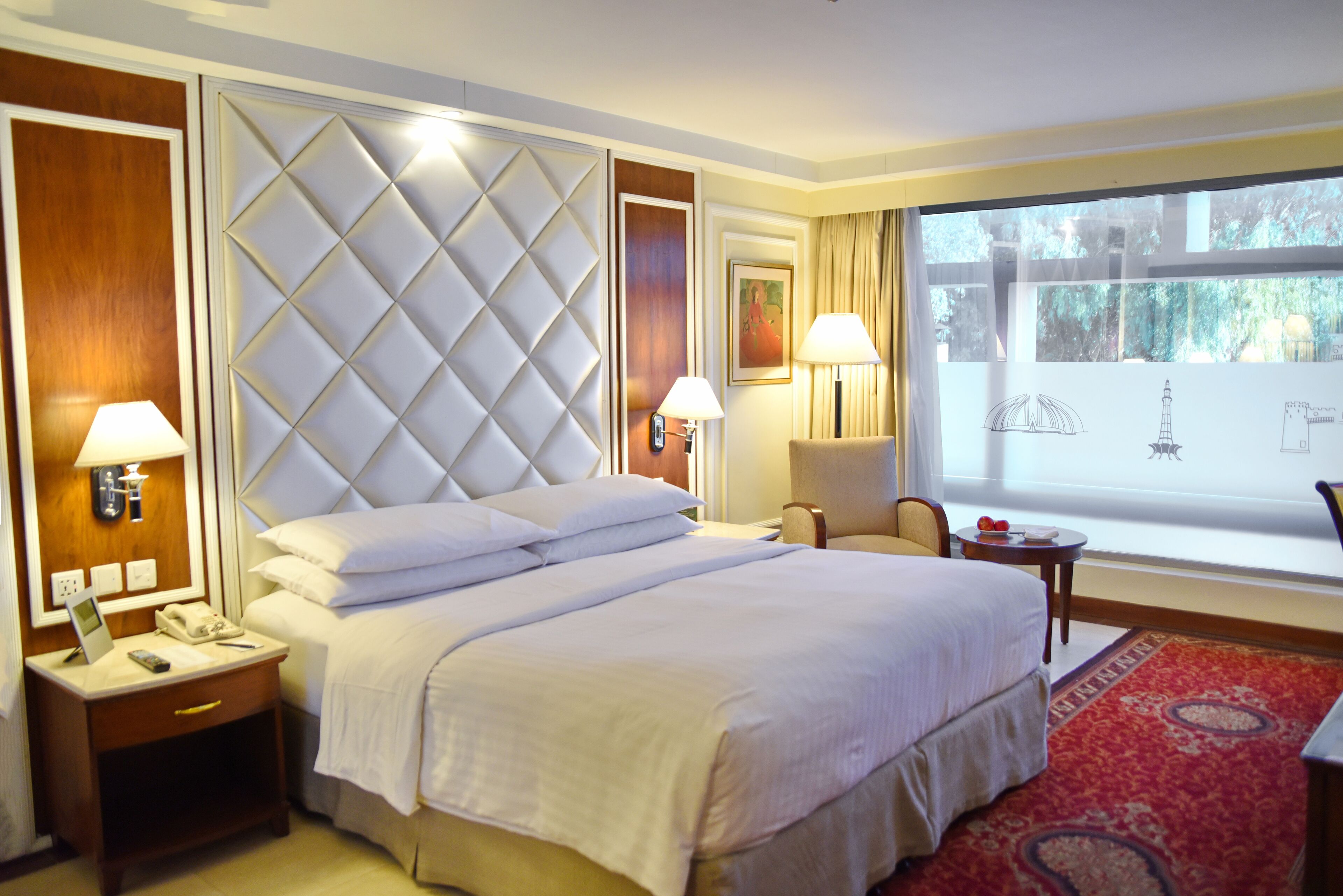 A hotel room with a large bed, two bedside tables, a chair, a window with a view, and a red carpet.