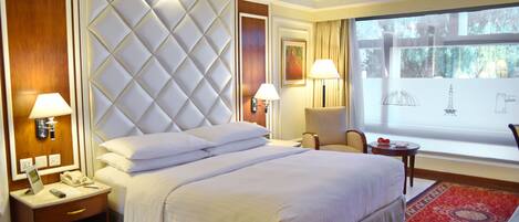 A hotel room with a large bed, two bedside tables, a chair, a window with a view, and a red carpet.