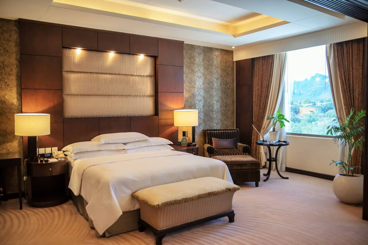 Una habitación de hotel con una cama grande, dos lámparas de noche, una silla, una mesita y una planta en maceta.