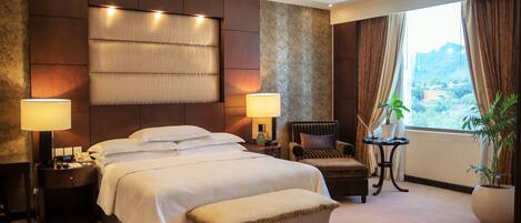 A hotel room with a large bed, two bedside lamps, a chair, a small table, and a potted plant.