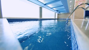 Una piscina interior con agua azul cristalina, una escalera metálica y un techo con vigas azules.