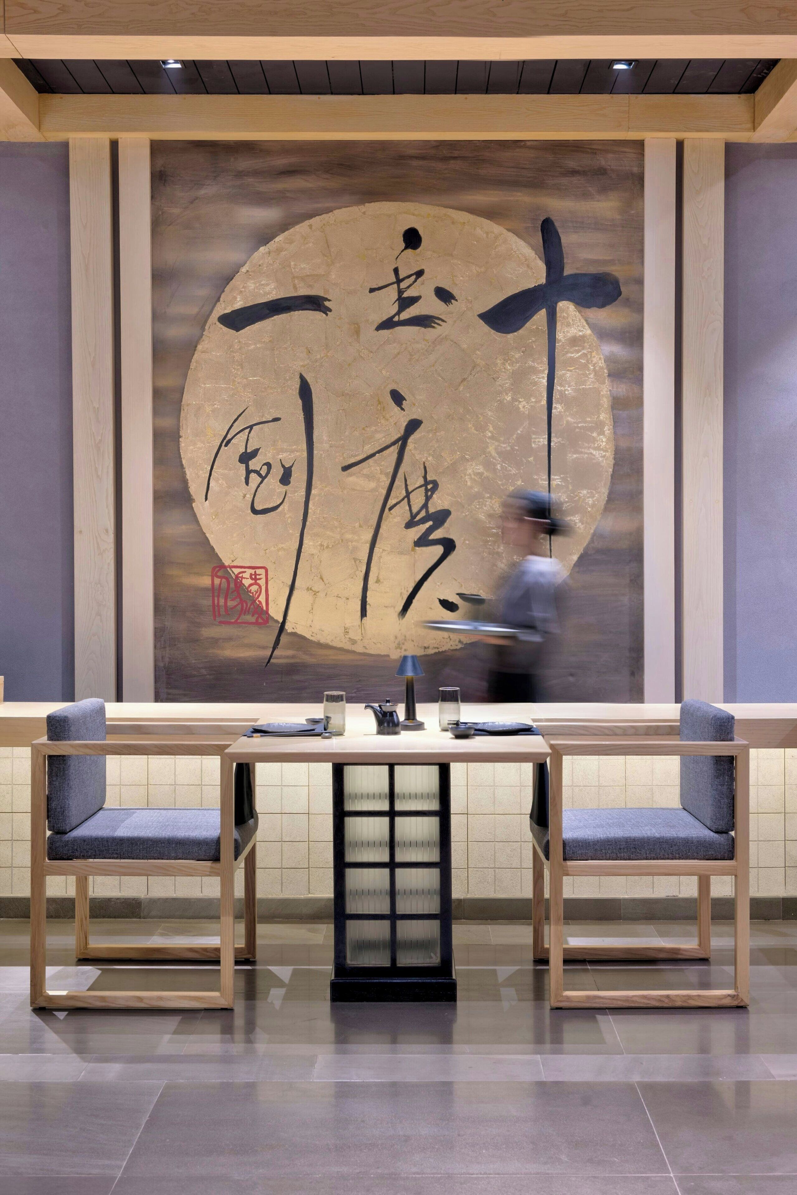 A restaurant interior with a large calligraphy artwork on the wall, wooden furniture, and a tiled floor.