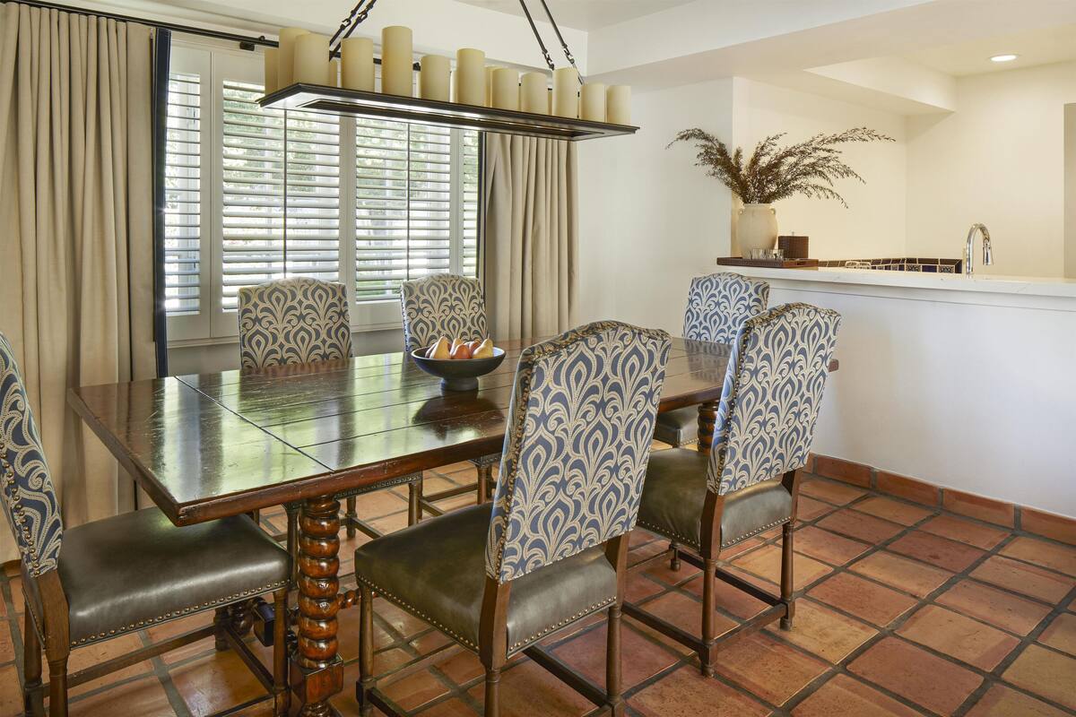 A dining area with a wooden table, patterned chairs, a bowl of fruit, and a kitchen in the background.