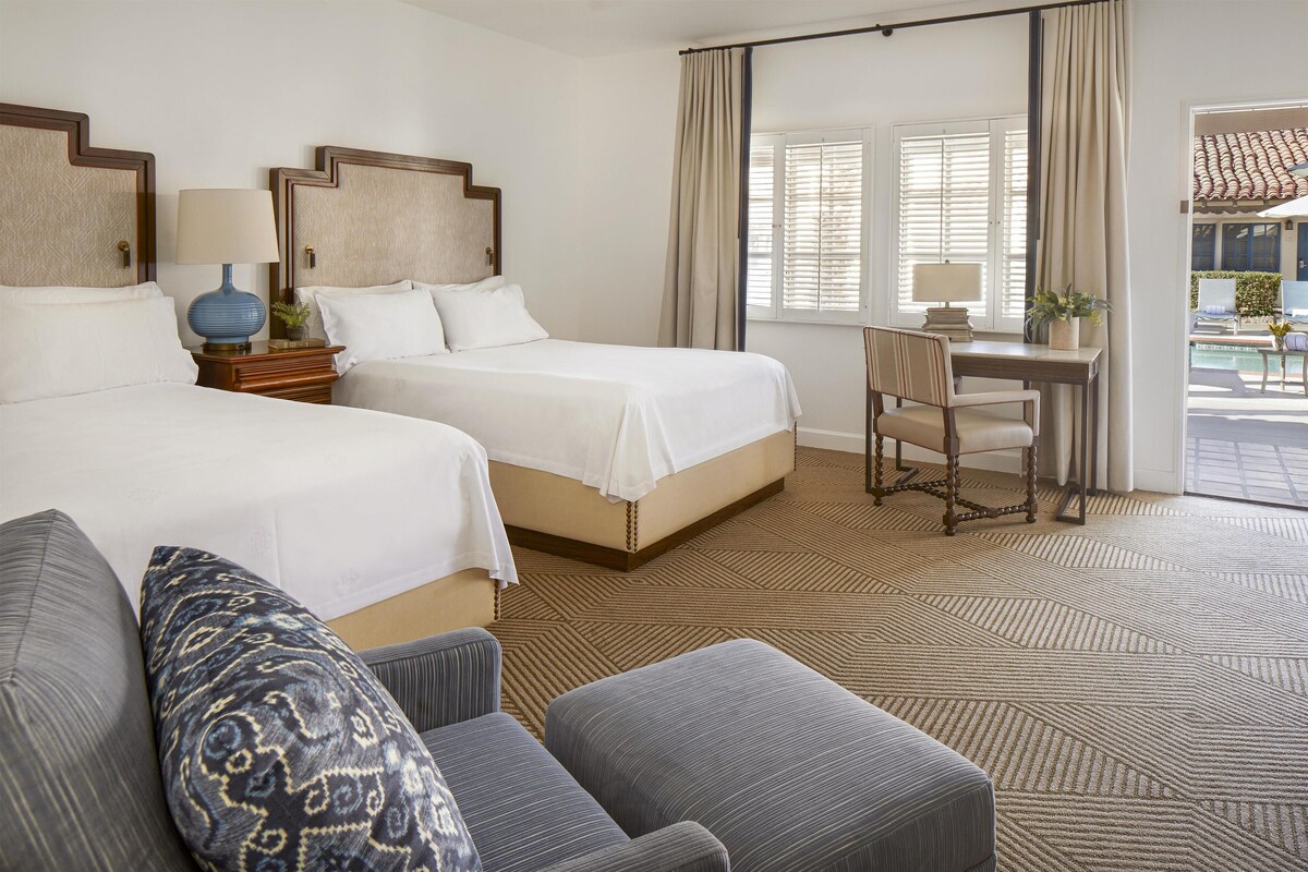 A hotel room with two beds, a desk, and a view of the outside.