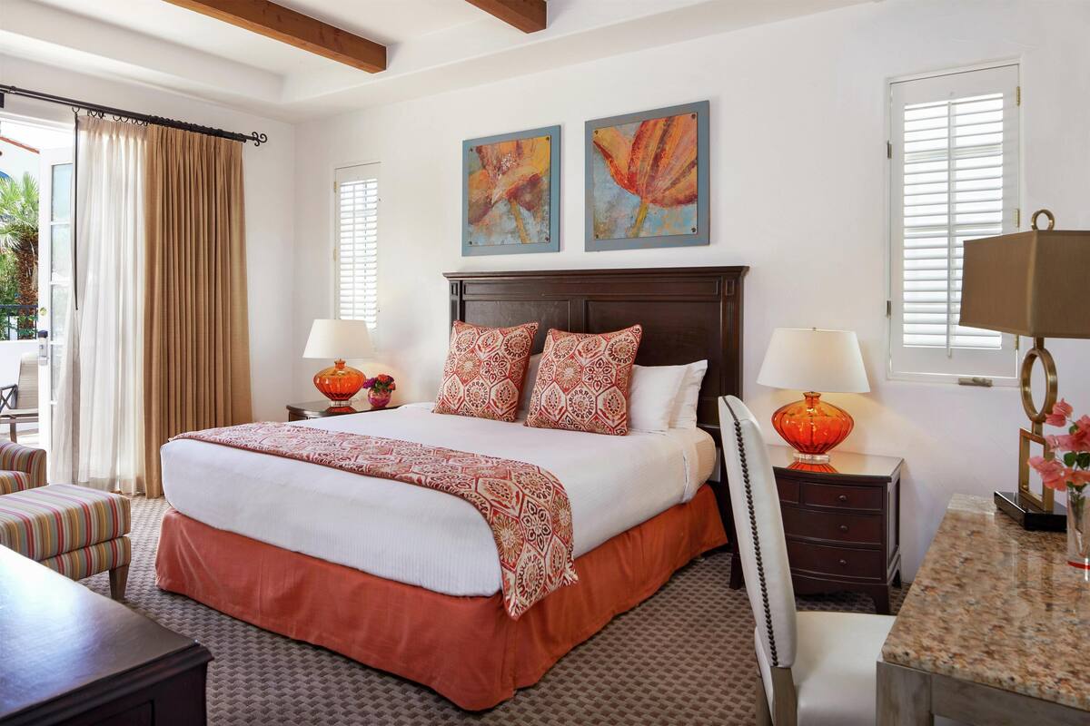 A hotel room with a large bed, two bedside tables with lamps, a desk, and a chair. There are two paintings on the wall above the bed.