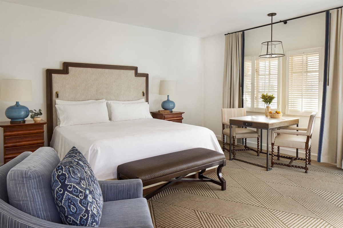 A hotel room with a large bed, a desk with two chairs, a bench, and a patterned carpet.
