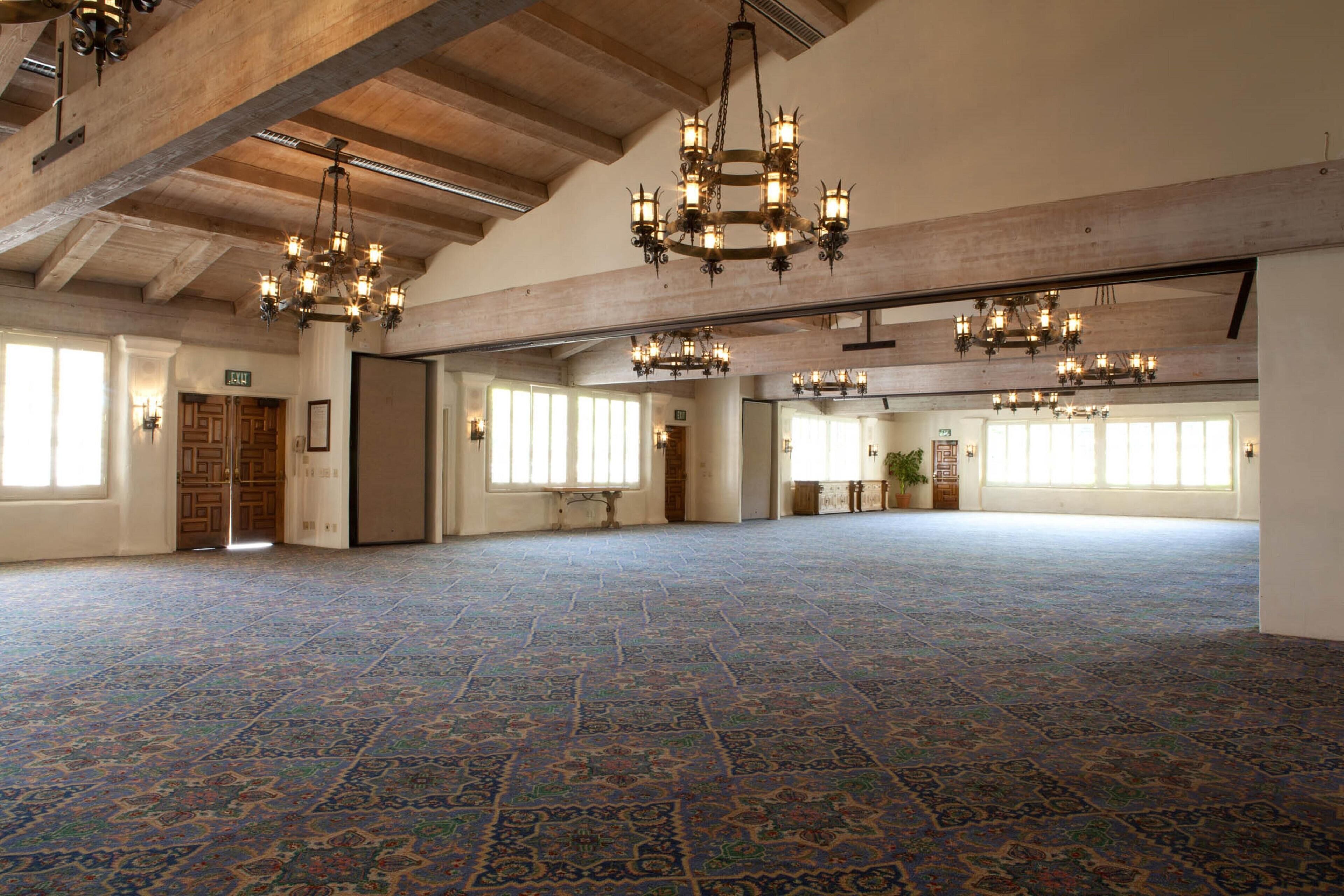 Un amplio salón con alfombra estampada, vigas de madera y múltiples candelabros.