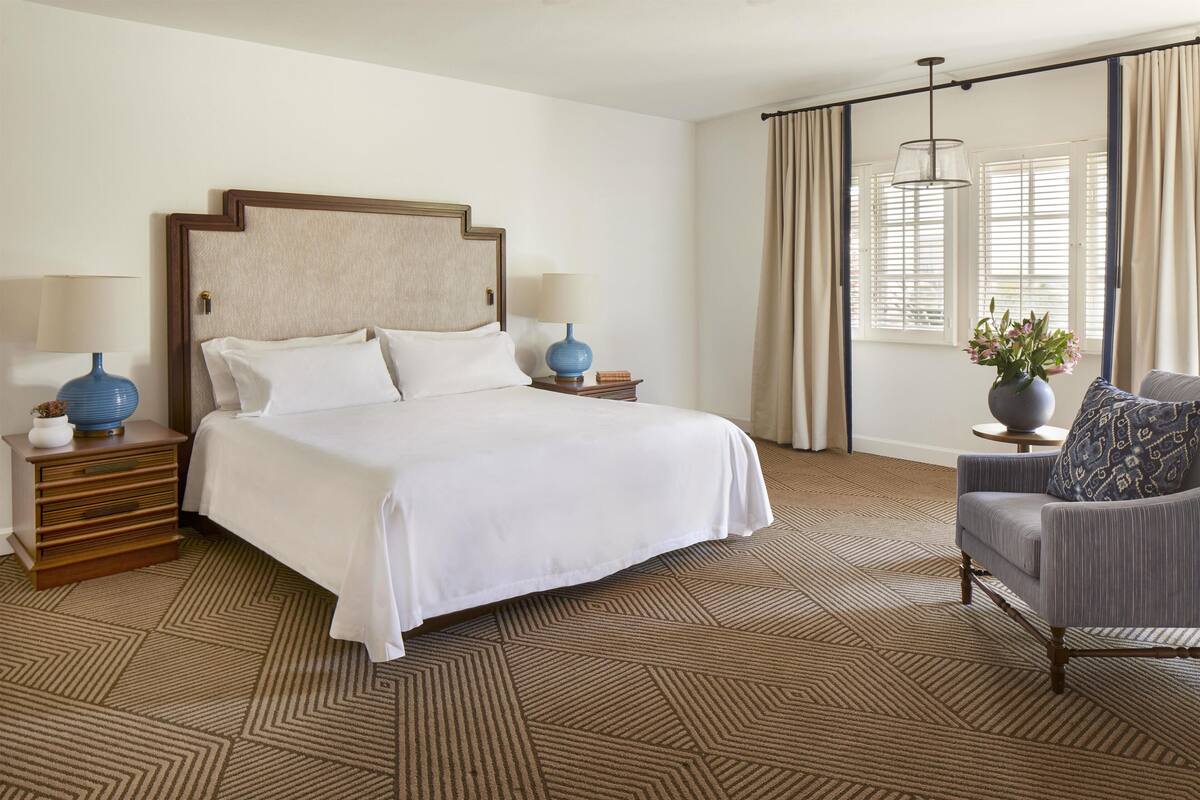 A hotel room with a large bed, two bedside tables with lamps, a chair, a potted plant, and a window with curtains.