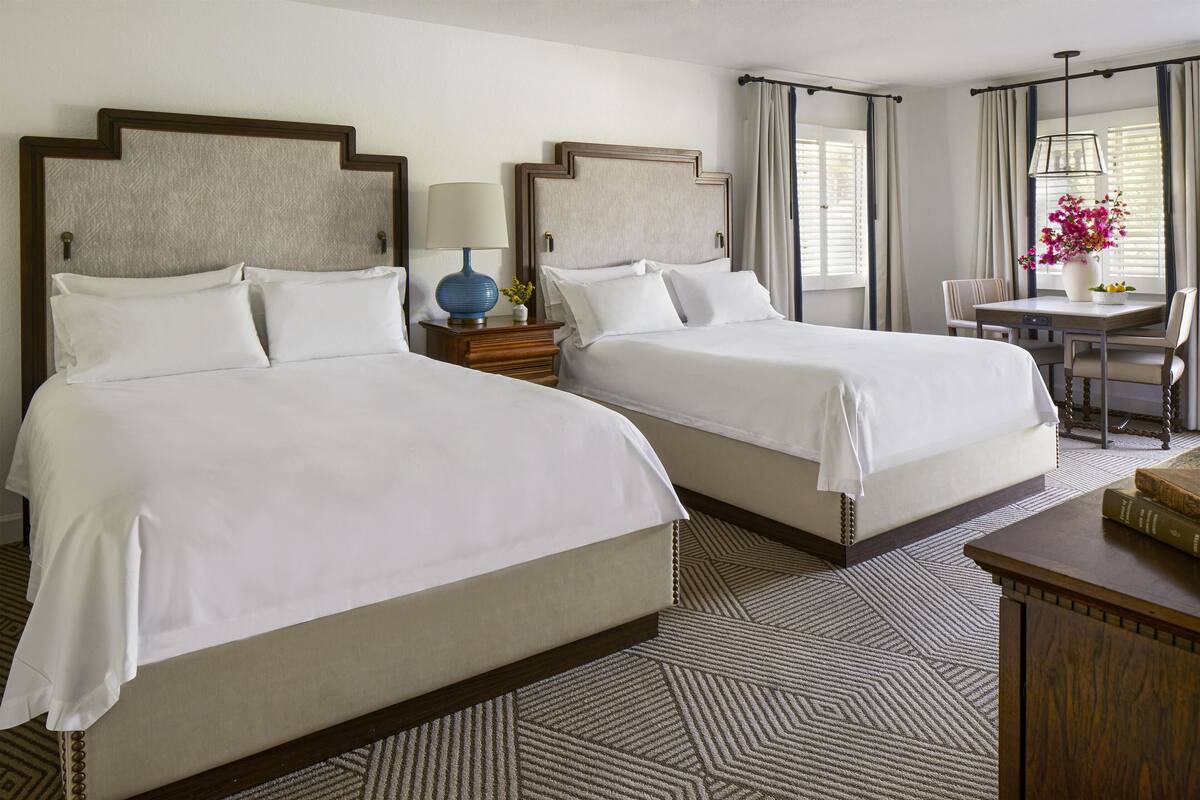 A hotel room with two beds, a small table with a vase of flowers, and a view of the outdoors through the window.
