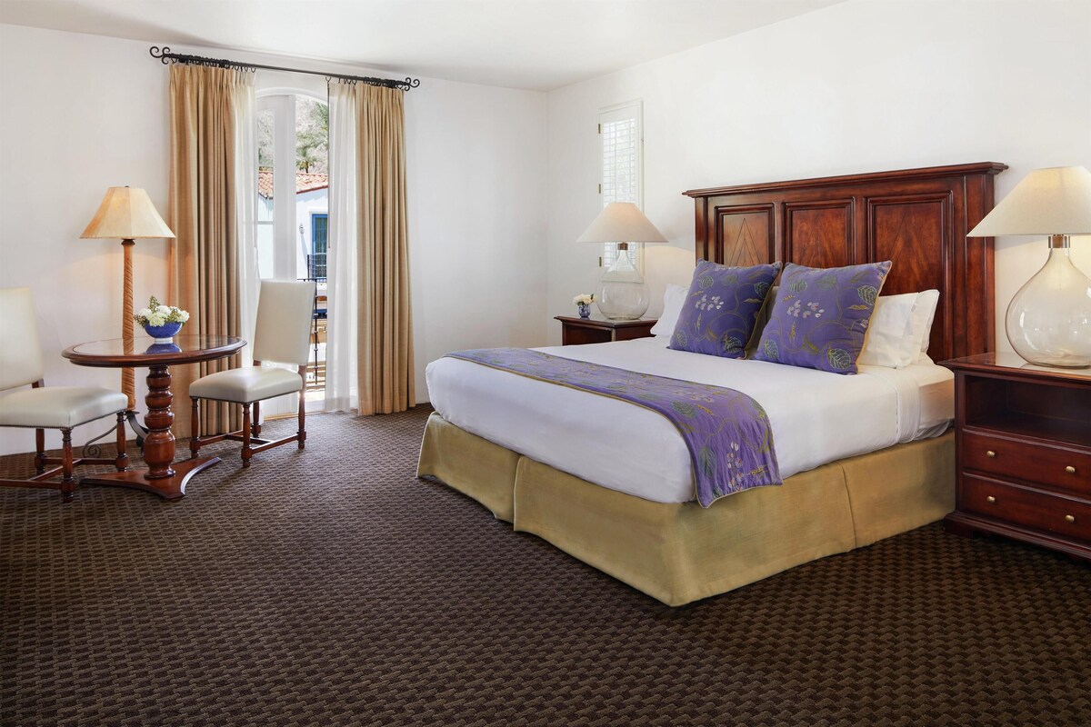 A hotel room with a large bed, two bedside tables with lamps, a chair, and a small table with a vase.
