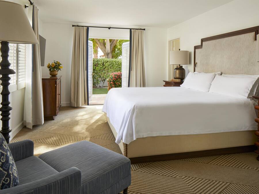 Quarto de hotel com uma cama grande, uma cadeira, uma mesa com luminária e vista para um jardim por uma porta aberta.