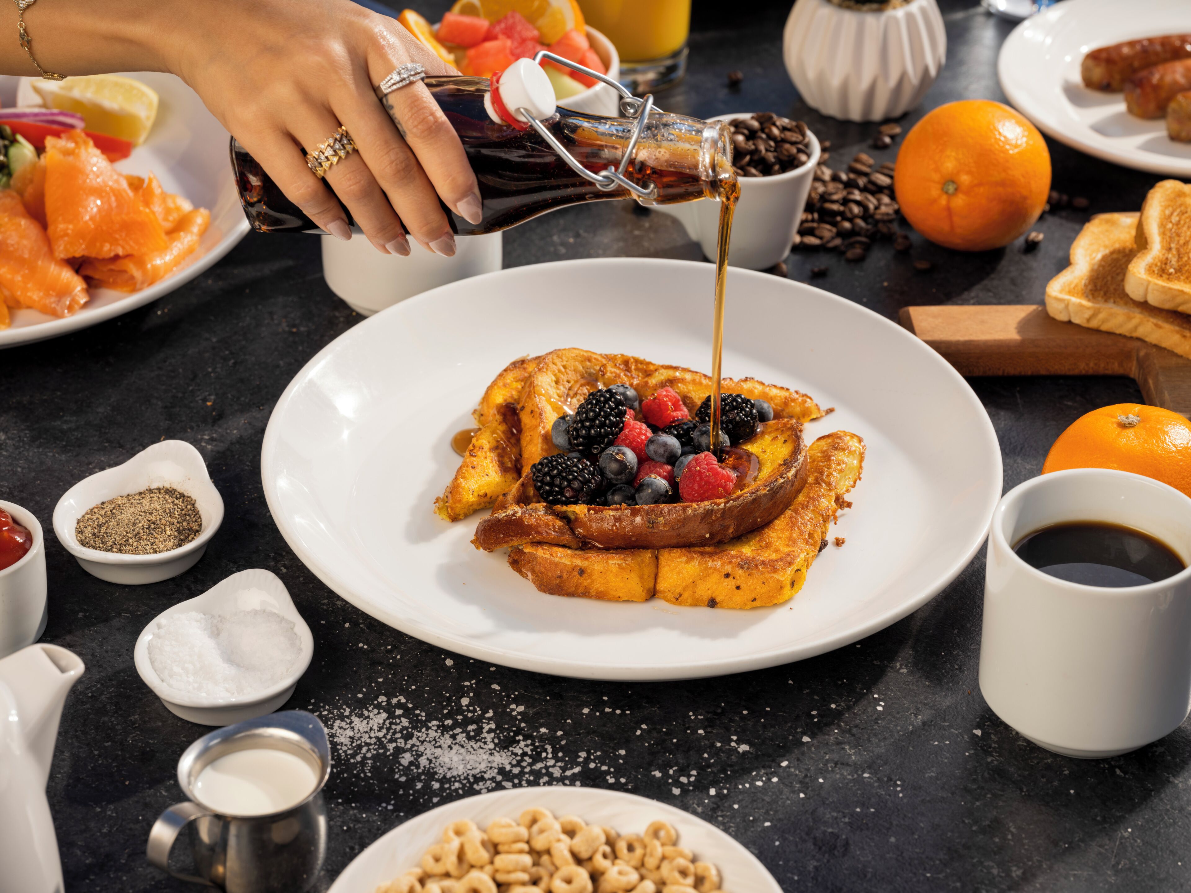 A breakfast plate with toast, berries, and syrup, surrounded by various food items and condiments on a table.