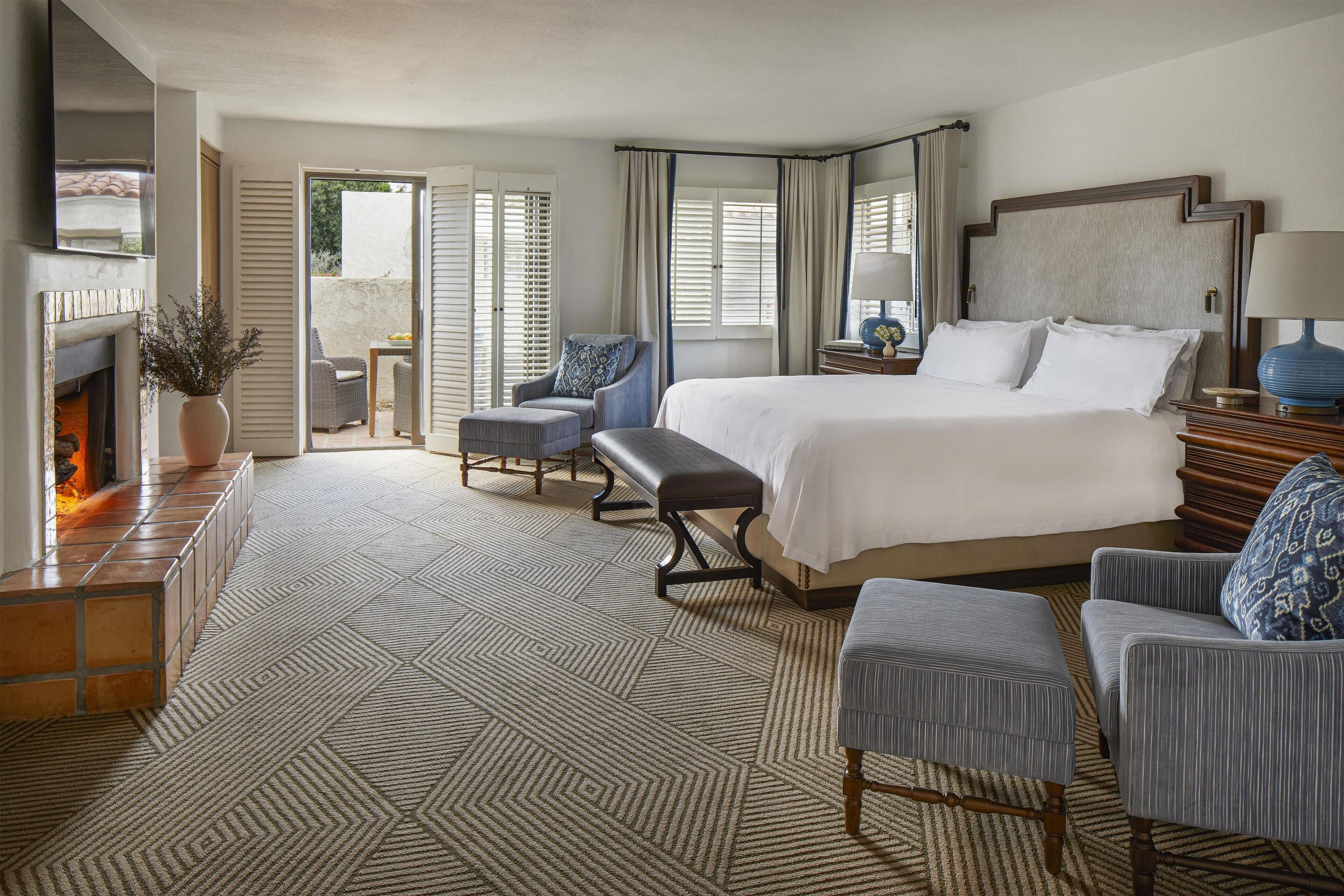 A hotel room with a large bed, two armchairs, a bench, a small table, and a fireplace.