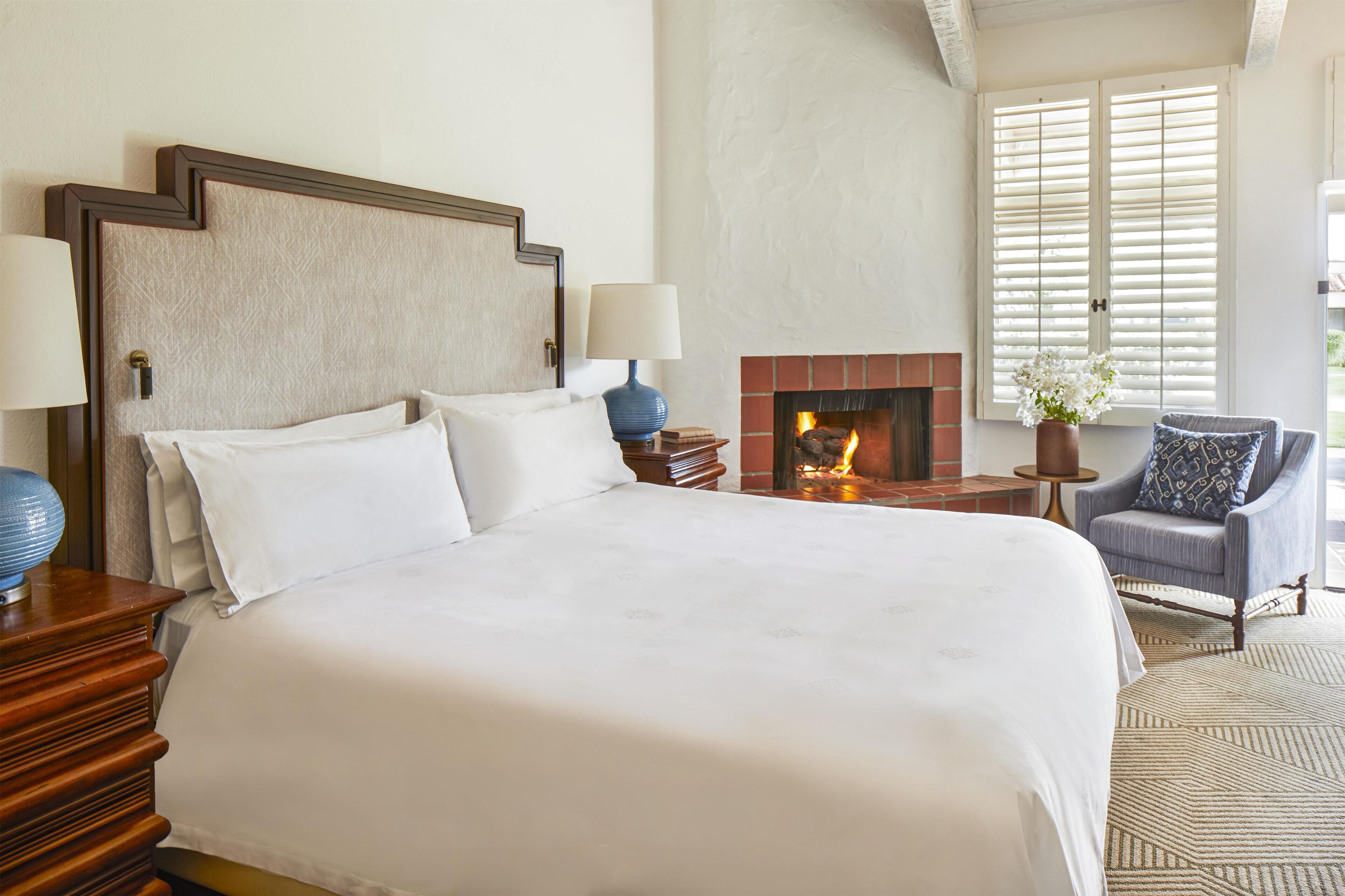 Una cama grande con cabecera de madera, dos lámparas de noche, una mesita con un florero, una silla con cojín estampado y una chimenea.