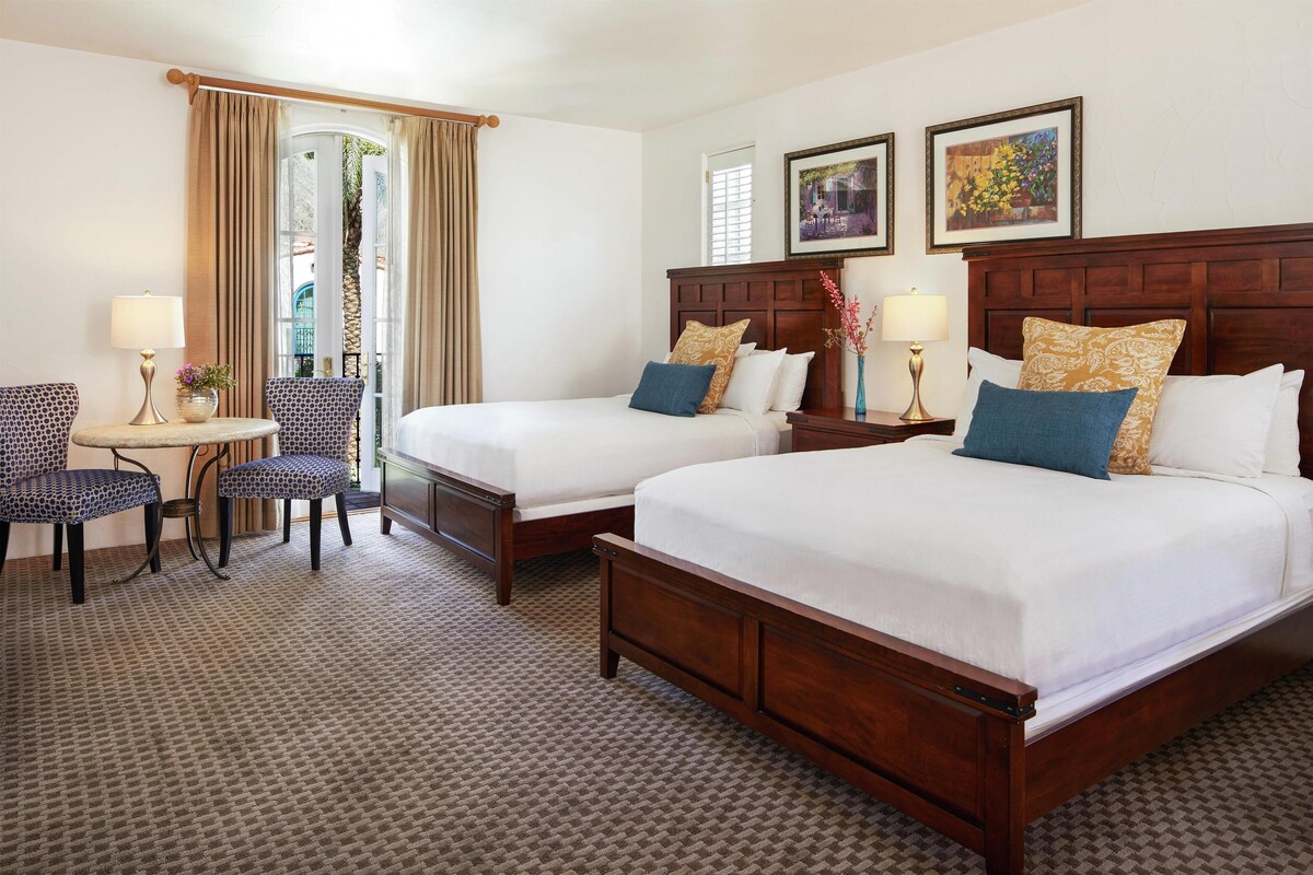 A hotel room with two beds, a sitting area with a table and chairs, and a view of the outdoors through a window.