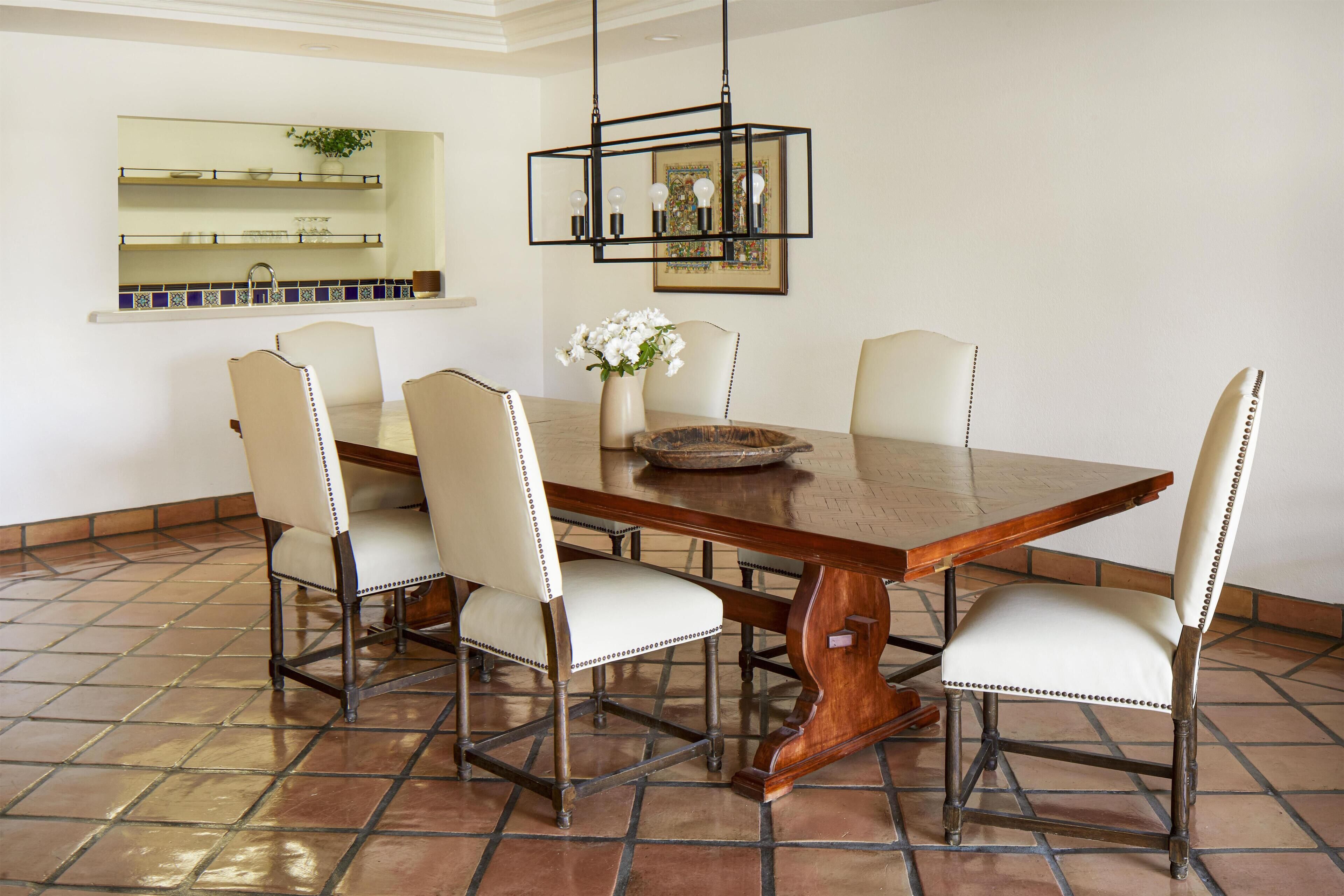 Un comedor con una gran mesa de madera, seis sillas, un florero con flores y una lámpara colgante.