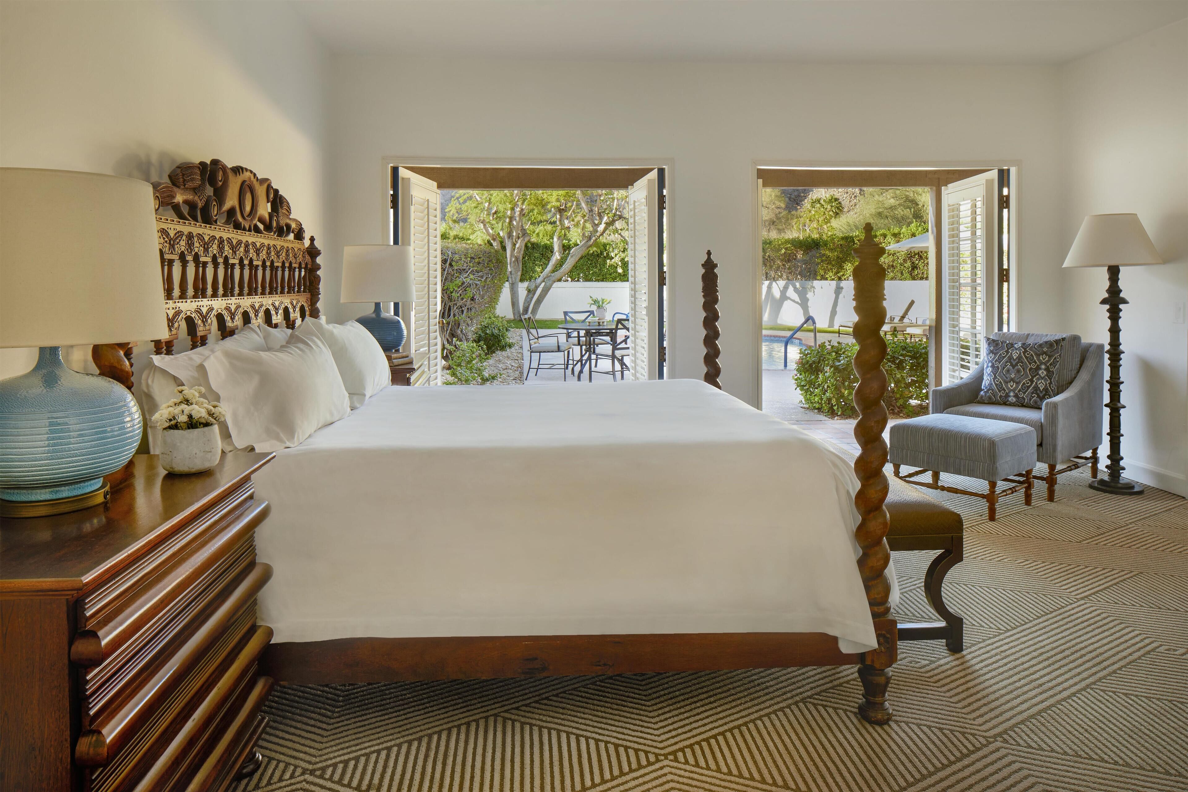 Une chambre dotée d’un grand lit, d’une tête de lit en bois, de deux tables de chevet avec des lampes, d’une chaise et donnant sur une terrasse extérieure avec une table et des chaises.