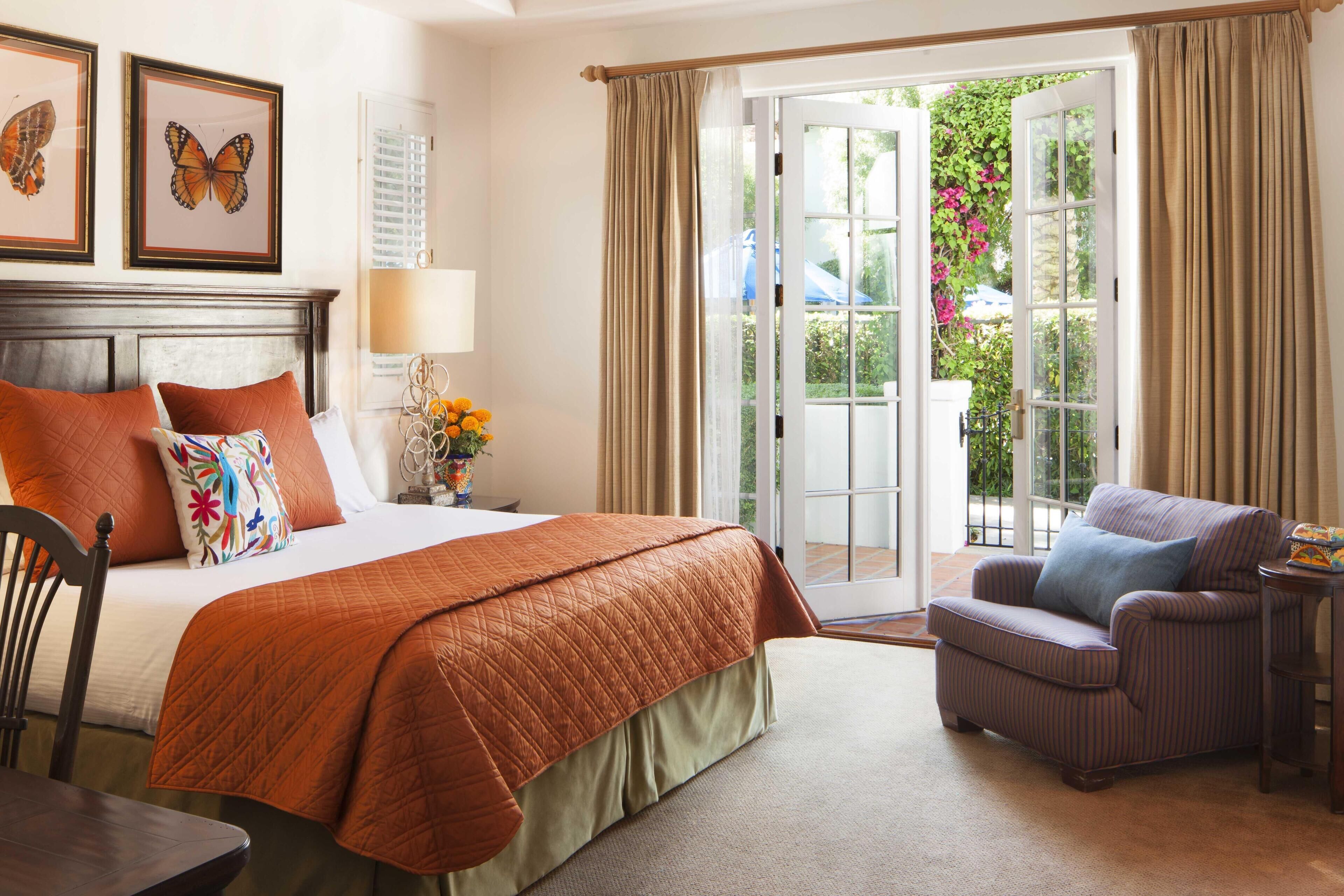 A hotel room with a large bed, a chair, a table, and a view of the garden through the door.