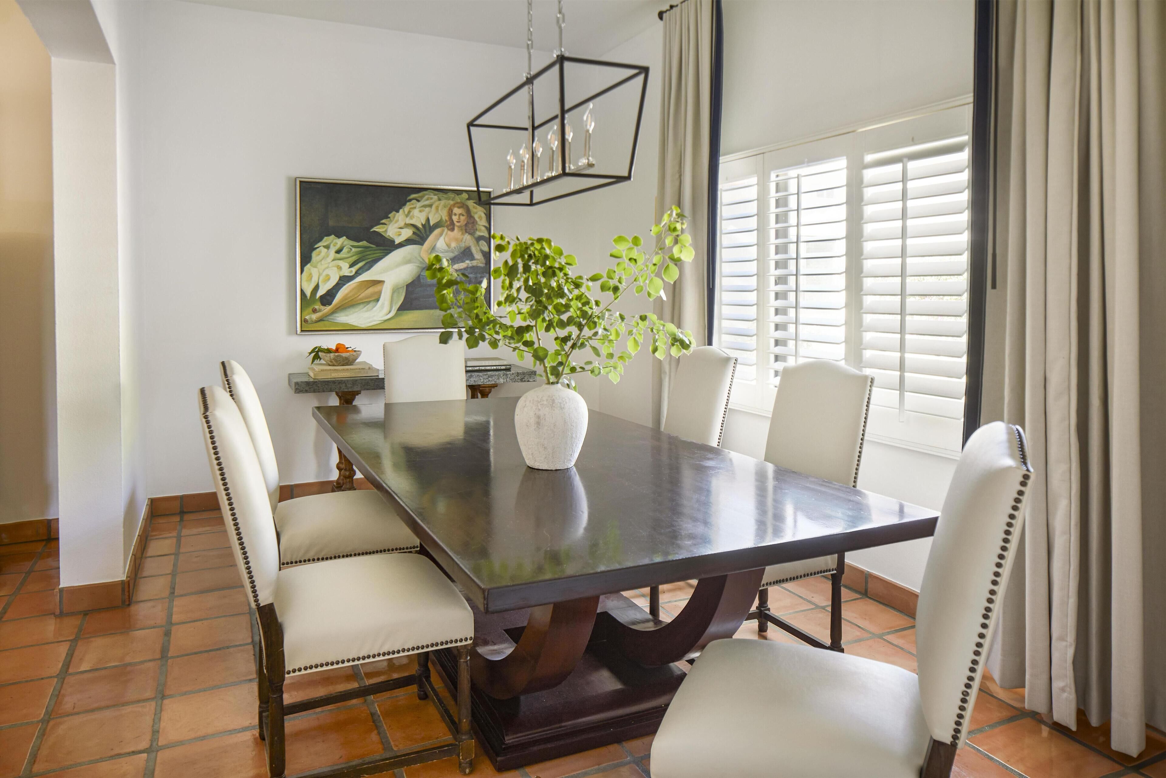A dining area with a rectangular table, six chairs, a vase with a plant, a painting on the wall, and a window with blinds.