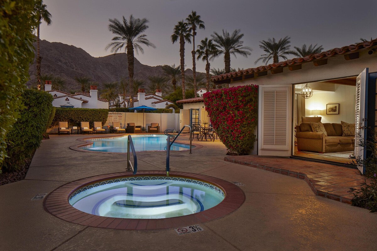 A pool area with hot tubs, a lounge chair, and a patio with a view of palm trees and a mountain.