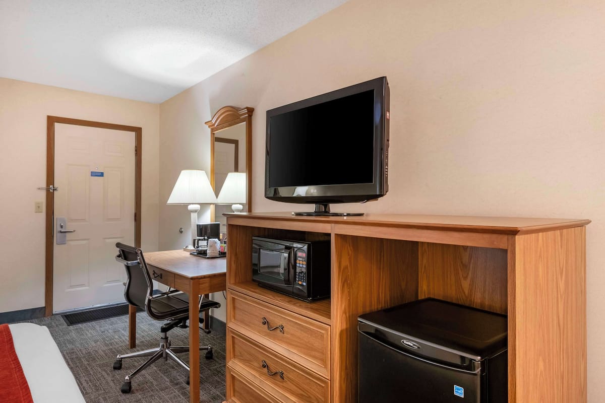 Ein Hotelzimmer mit Holzschreibtisch, Fernseher, Mikrowelle, Kühlschrank, Stuhl und einem Bett mit roter Decke.