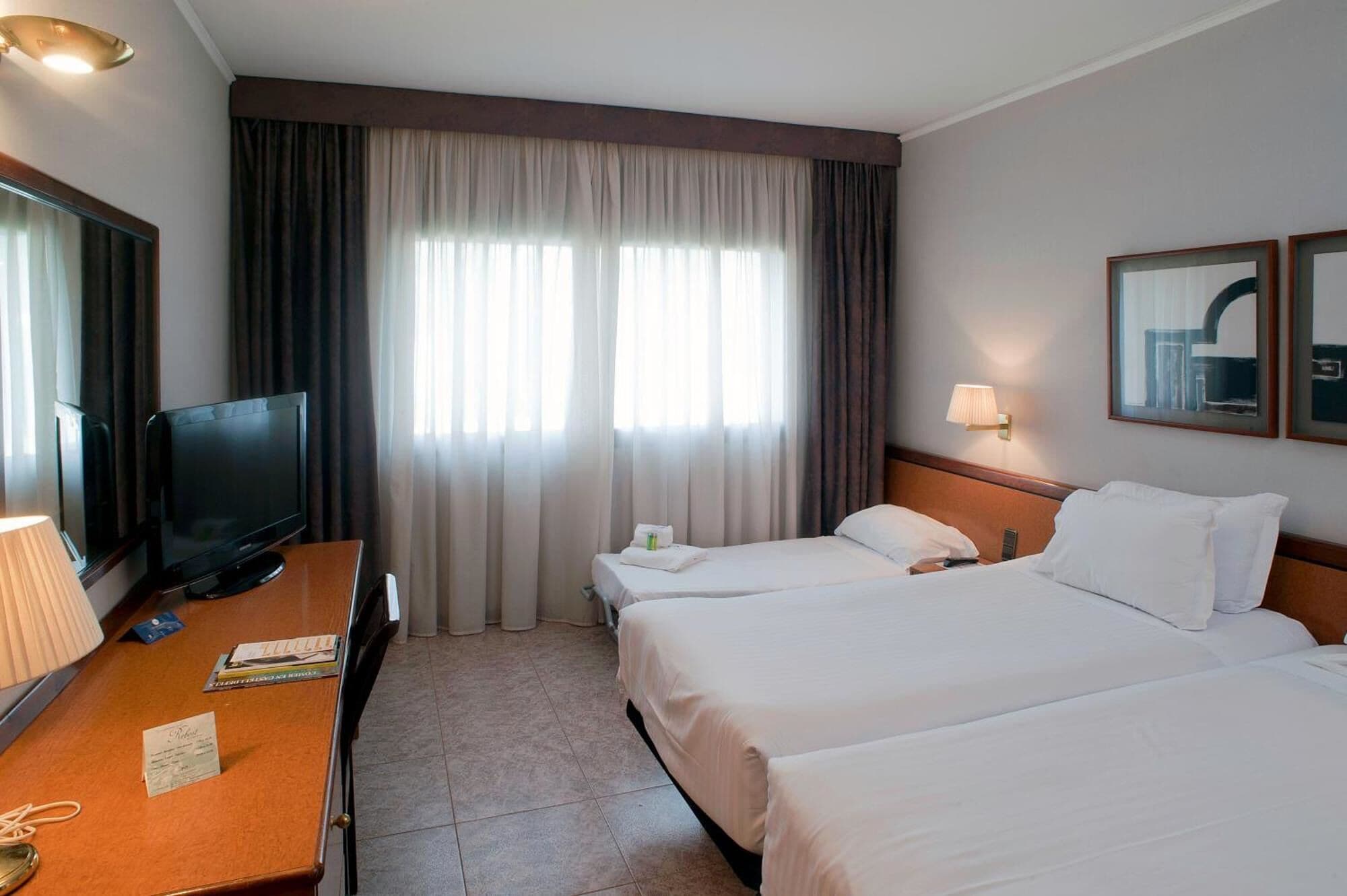 Habitación de hotel con televisión, escritorio, dos camas, ventana con cortinas y una lámpara de pared.