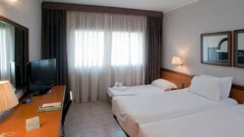 A hotel room with a TV, a desk, two beds, a window with curtains, and a wall-mounted lamp.