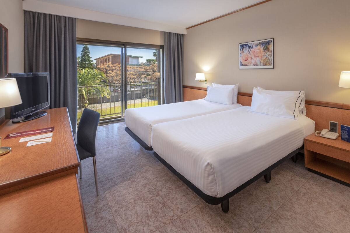 Une chambre d’hôtel avec deux lits, un bureau avec une télévision, un téléphone et une fenêtre donnant sur un immeuble et des arbres.