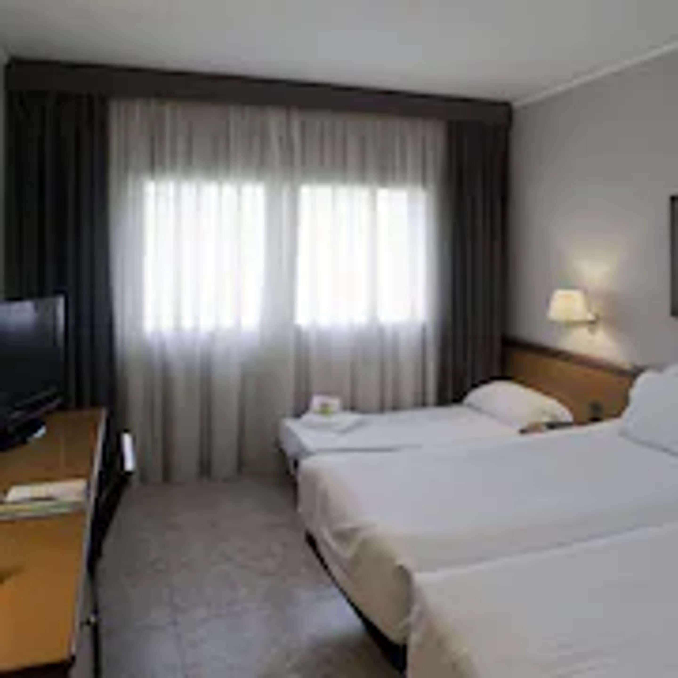 A hotel room with two beds, a TV, a desk, and a window with curtains.