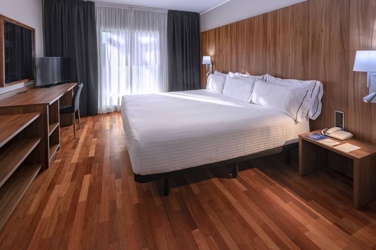 Une chambre d’hôtel avec un plancher en bois, un lit avec du linge de maison blanc, un bureau avec une télévision, une table de chevet avec une lampe et une fenêtre avec des rideaux.